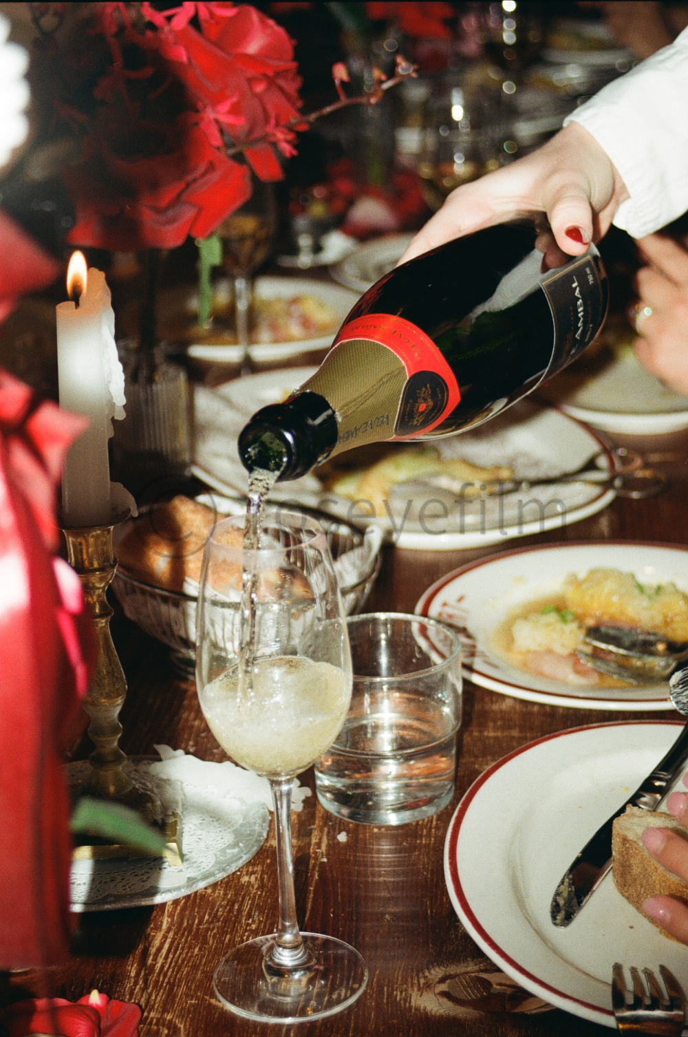 A person pours champagne into a glass at a festive dinner table with plates of food, a candle, and holiday decorations.