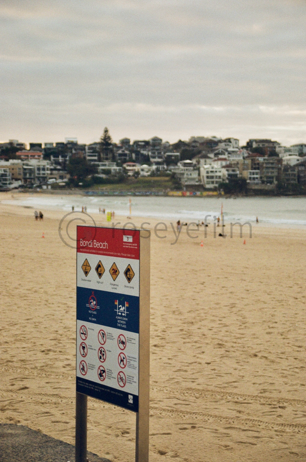 A beach scene at Bondi Beach with a warning sign in the foreground, sandy shore, ocean waves, and residential buildings on a hillside in the background, under a cloudy sky.