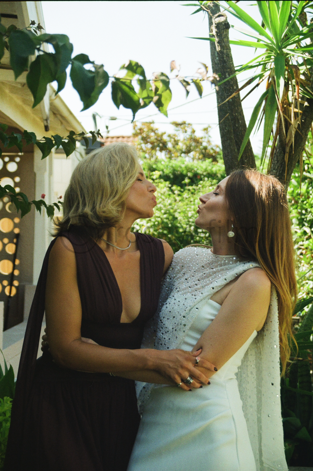 Two women are outdoors, standing close and about to kiss. One has gray hair and is wearing a dark sleeveless dress, and the other has long red hair and is wearing a white, beaded dress. There are trees and plants around them.