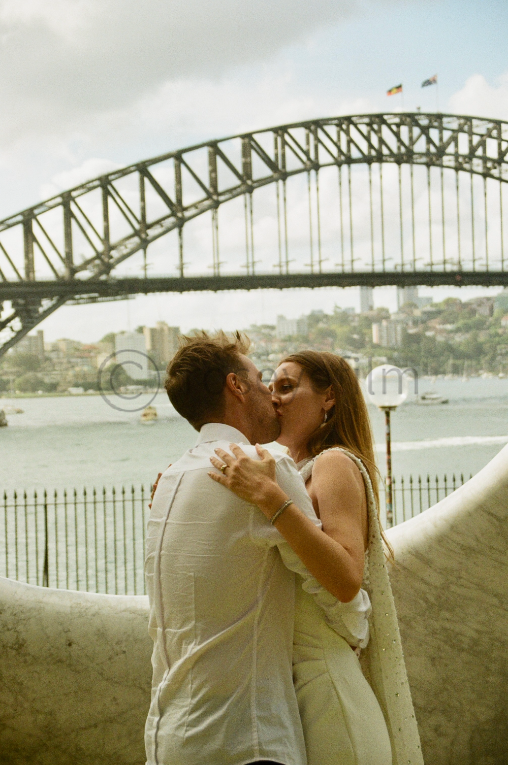 A couple kissing by the Sydney Harbour Bridge with water and buildings in the background.