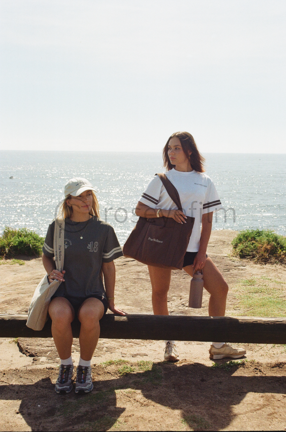 Two women near the ocean, one sitting on a wooden log wearing a dark t-shirt, shorts, a hat, and sneakers, the other standing on the ground with one leg raised on the log, holding a water bottle, wearing a white t-shirt, shorts, and sneakers, with la