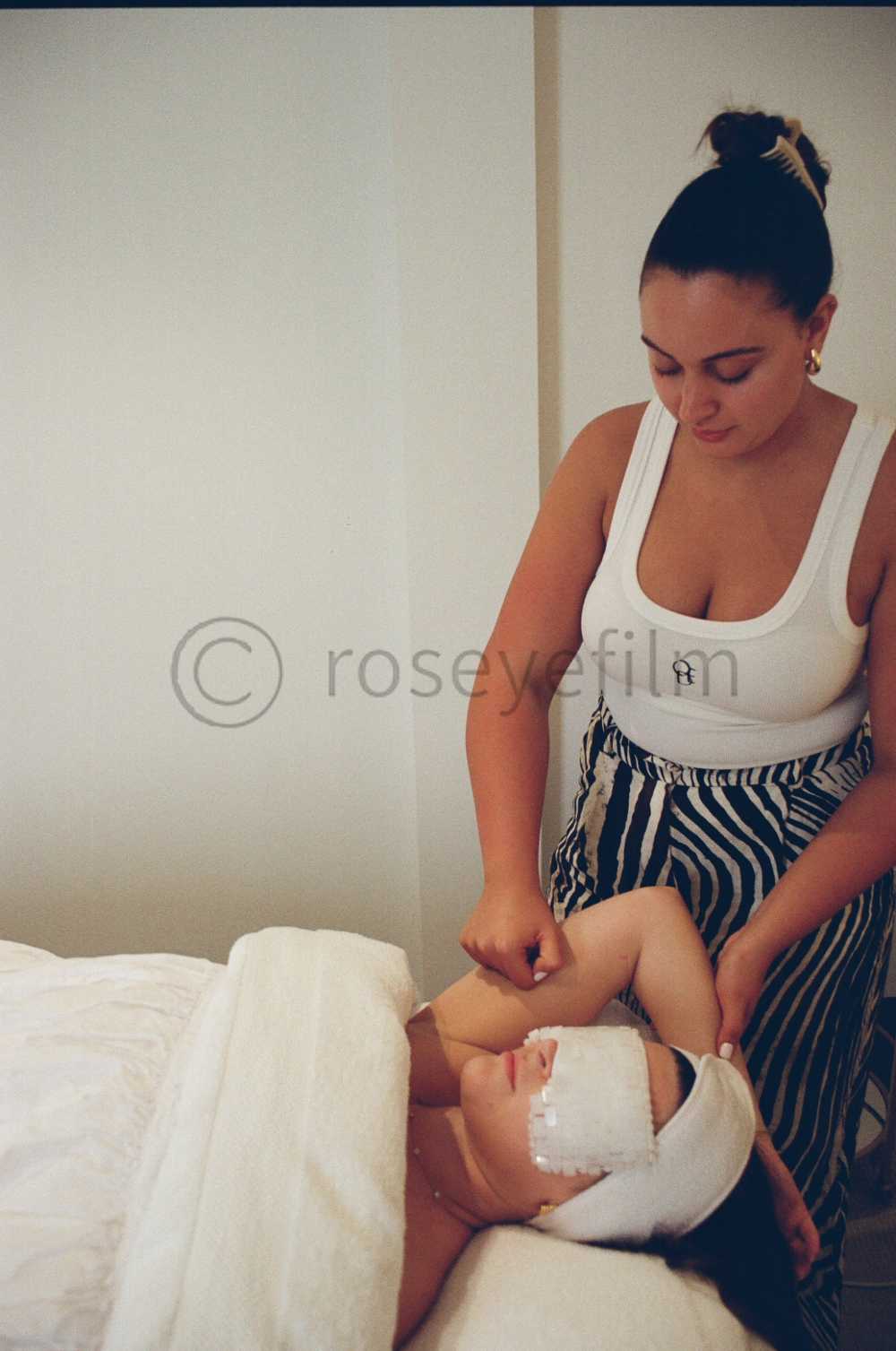 Woman in striped pants giving a massage to a woman lying on a bed with a white towel over her face and a nose strip, in a room with plain beige walls.