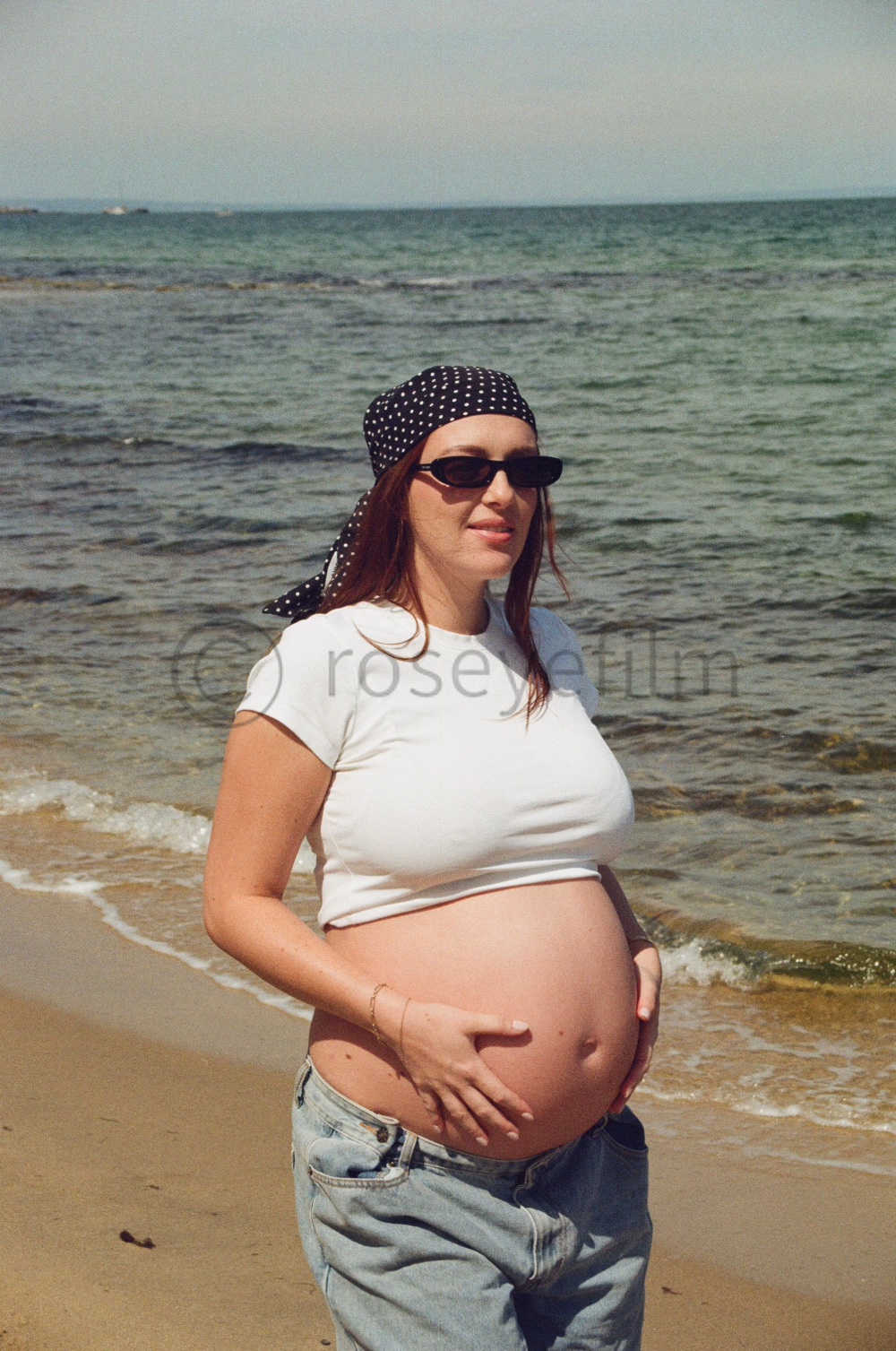 A pregnant woman standing on the beach near the ocean, wearing sunglasses, a black polka dot headscarf, a white crop top, and light blue jeans.