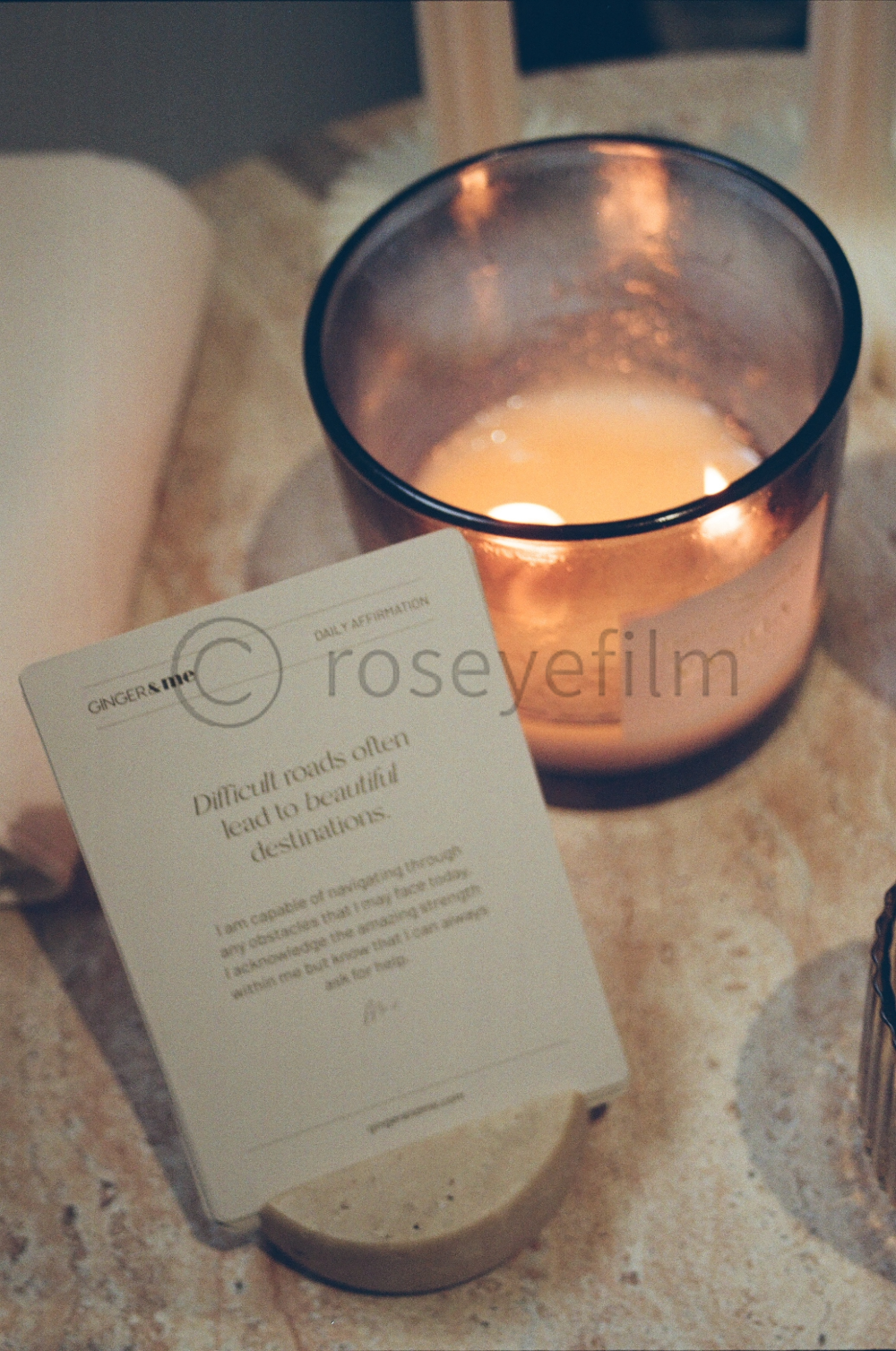 A lit candle in a glass holder next to a card with an affirmation about difficulty leading to beautiful destinations.