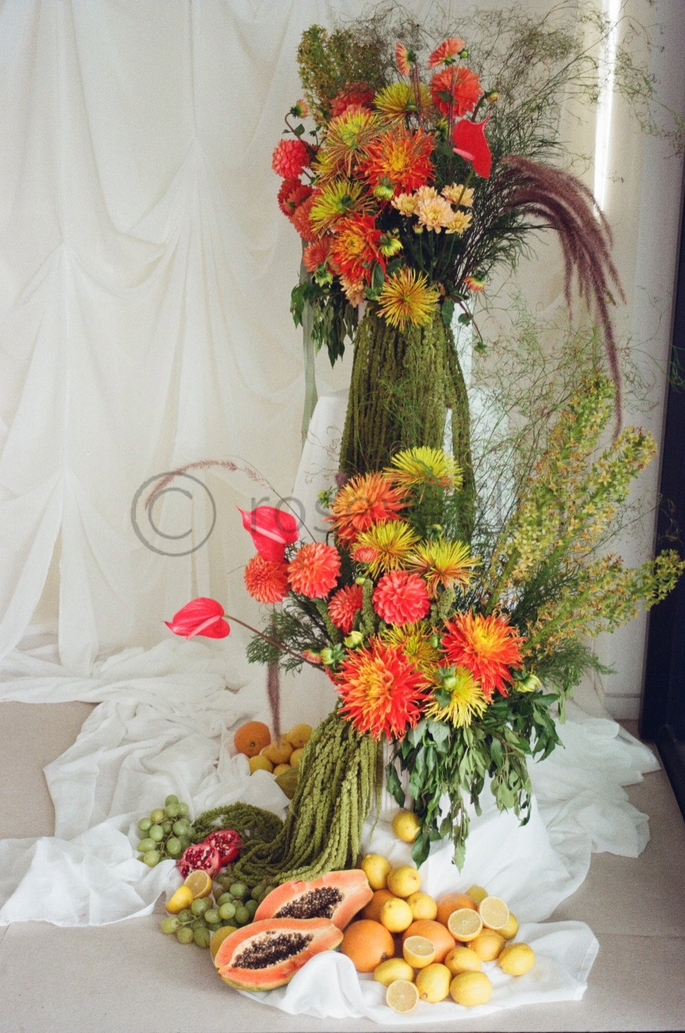 A large floral arrangement with orange and yellow flowers, green foliage, and tall, wispy grasses, displayed on white draped fabric with assorted fruits like papaya, lemons, grapes, and peaches at the base.