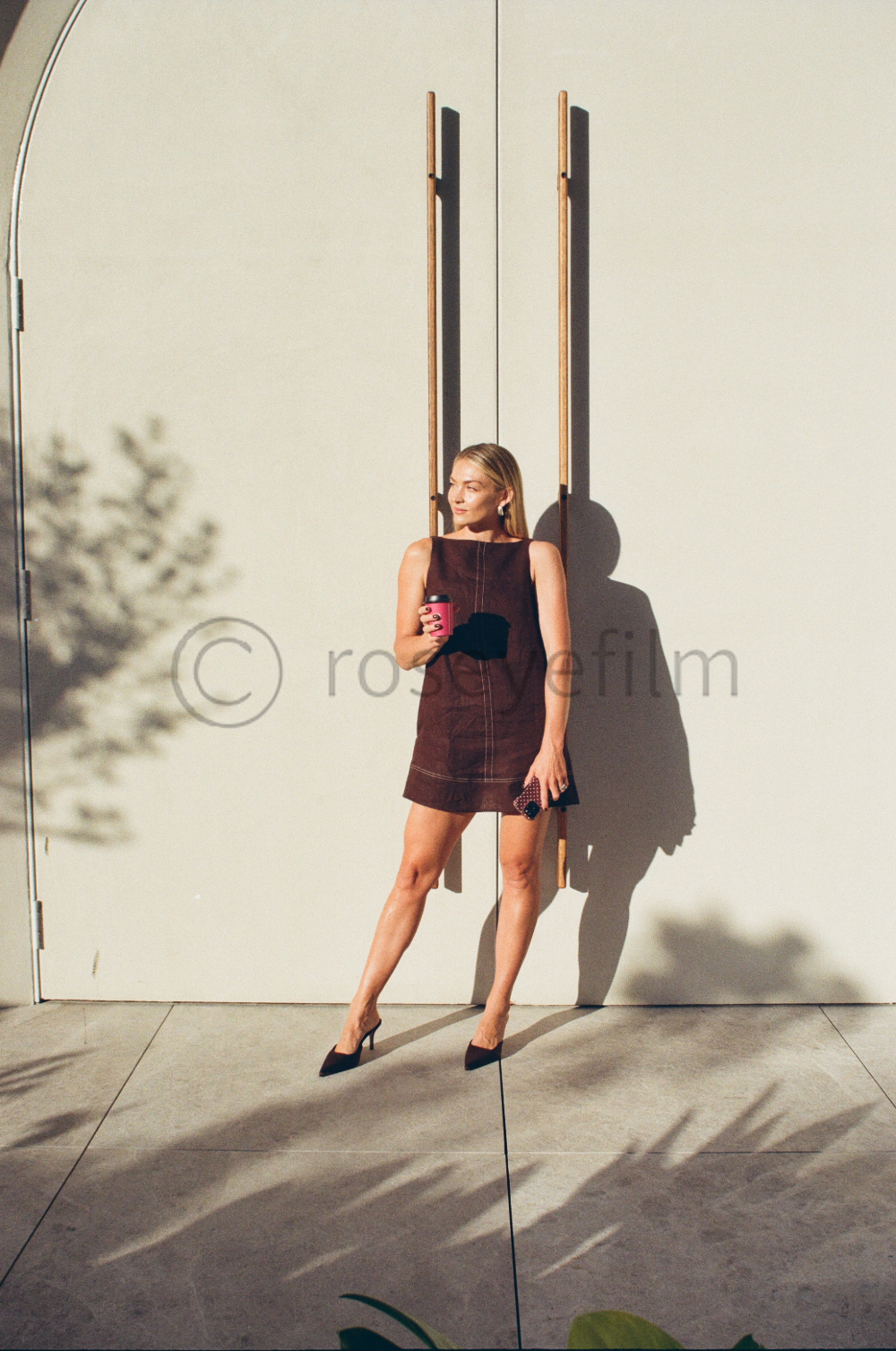 Woman in a sleeveless brown dress with high heels holding a pink coffee cup and a black phone, standing against a white wall with shadow of a tree on it.