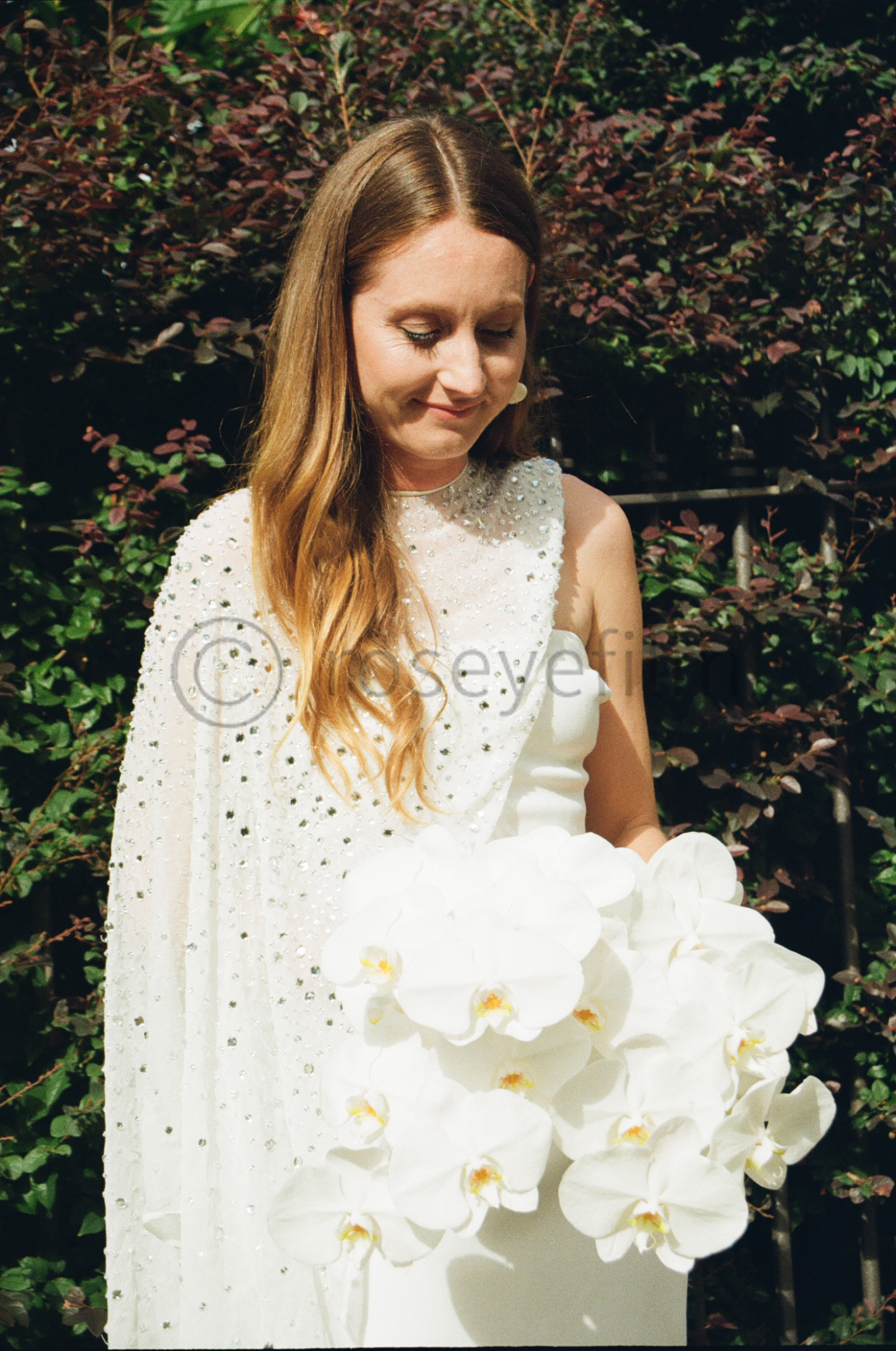 A woman with long, wavy hair wearing a white dress with decorative embellishments, standing outdoors in front of a leafy background, holding a large bouquet of white orchids.