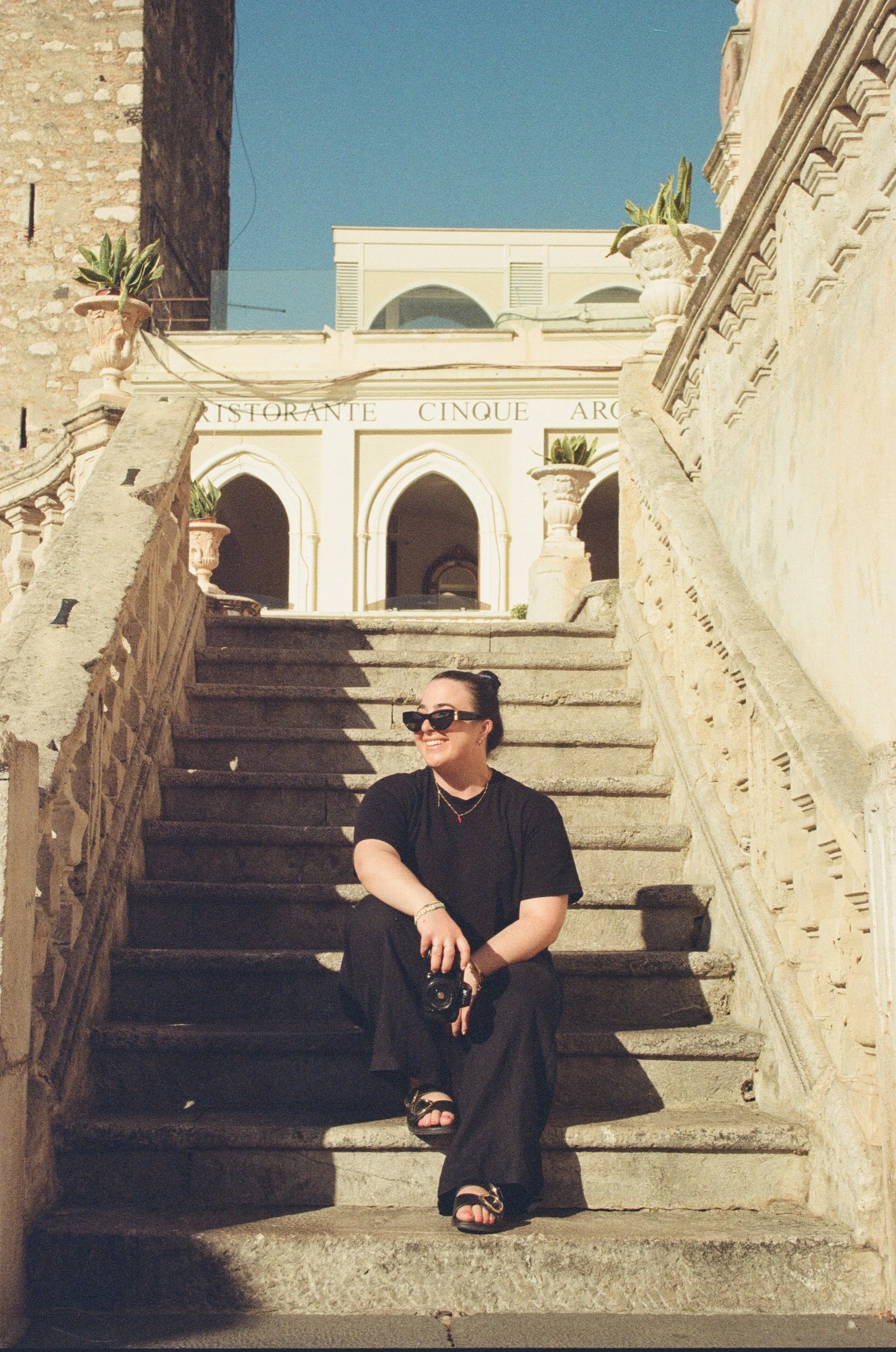 A woman sitting on granite stairs outside a historical building, wearing black clothing, sunglasses, and sandals, smiling and holding a camera.