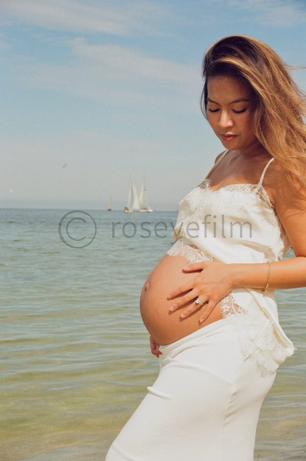 Pregnant woman in white dress standing by the sea, gently touching her baby bump, with sailboats in the background.