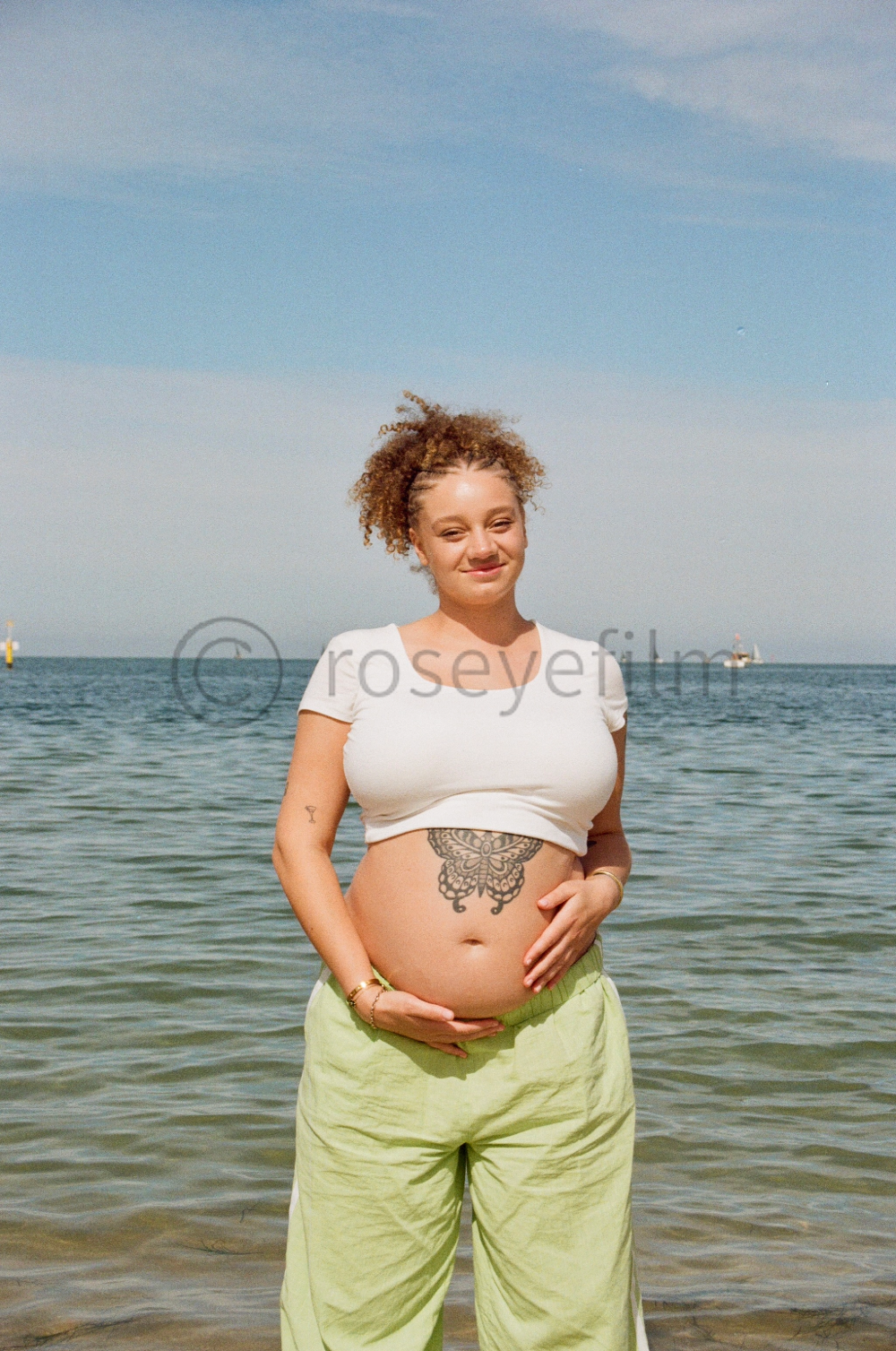 Pregnant woman standing in shallow ocean water, smiling, wearing a white crop top and light green pants, with a butterfly tattoo on her stomach, and tattoos on her left arm, holding her belly with both hands, under a blue sky.
