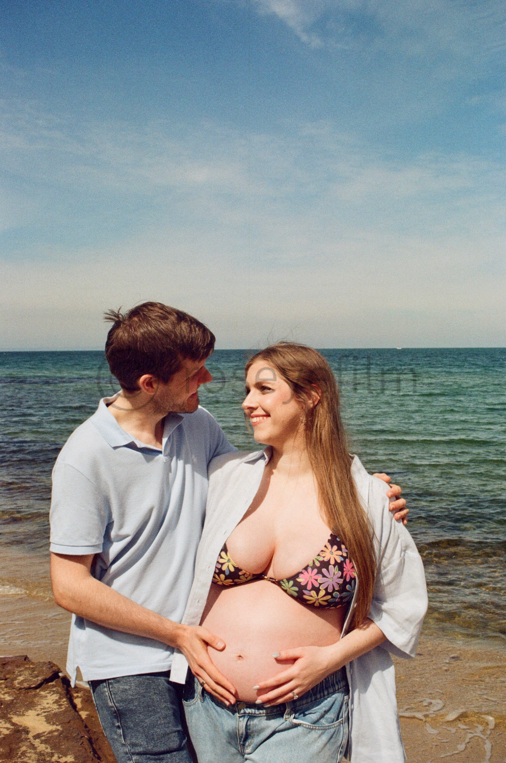 A pregnant woman and a man standing close by the beach, smiling at each other, with the ocean and blue sky in the background.