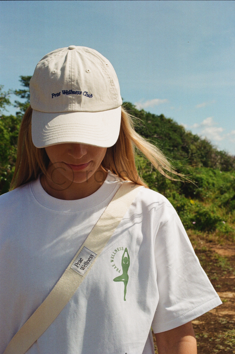 Woman wearing a white cap and t-shirt with 'Proe Wellness' branding, outdoors with greenery and a blue sky in the background.
