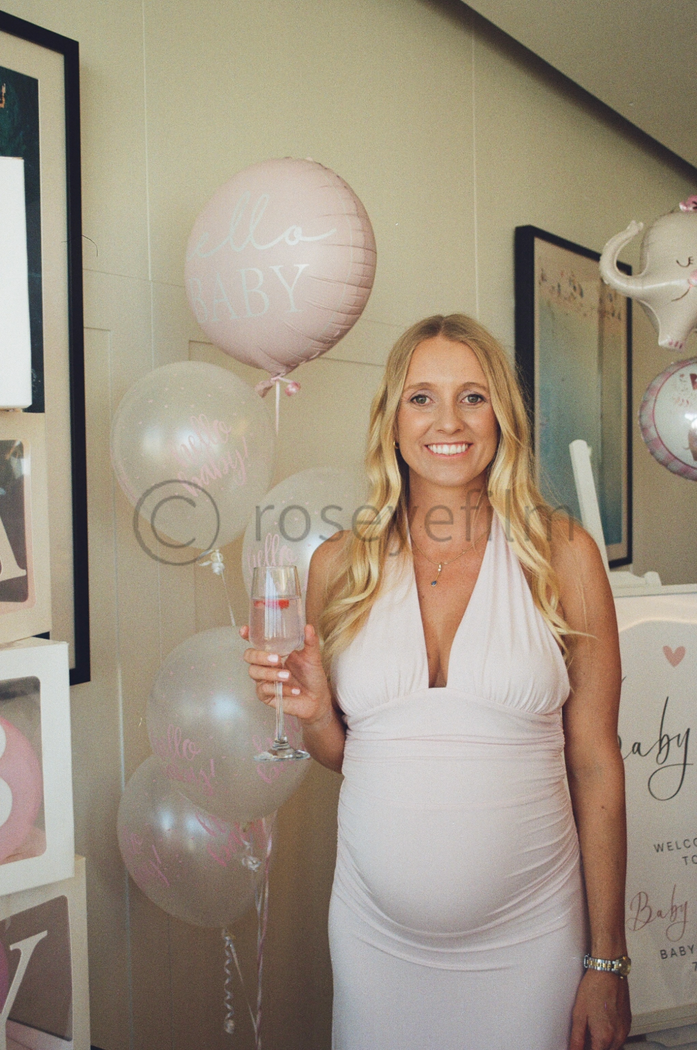A smiling woman at a baby shower holding a glass of champagne, surrounded by birthday balloons with messages like "Hello Baby" in a decorated indoor space.