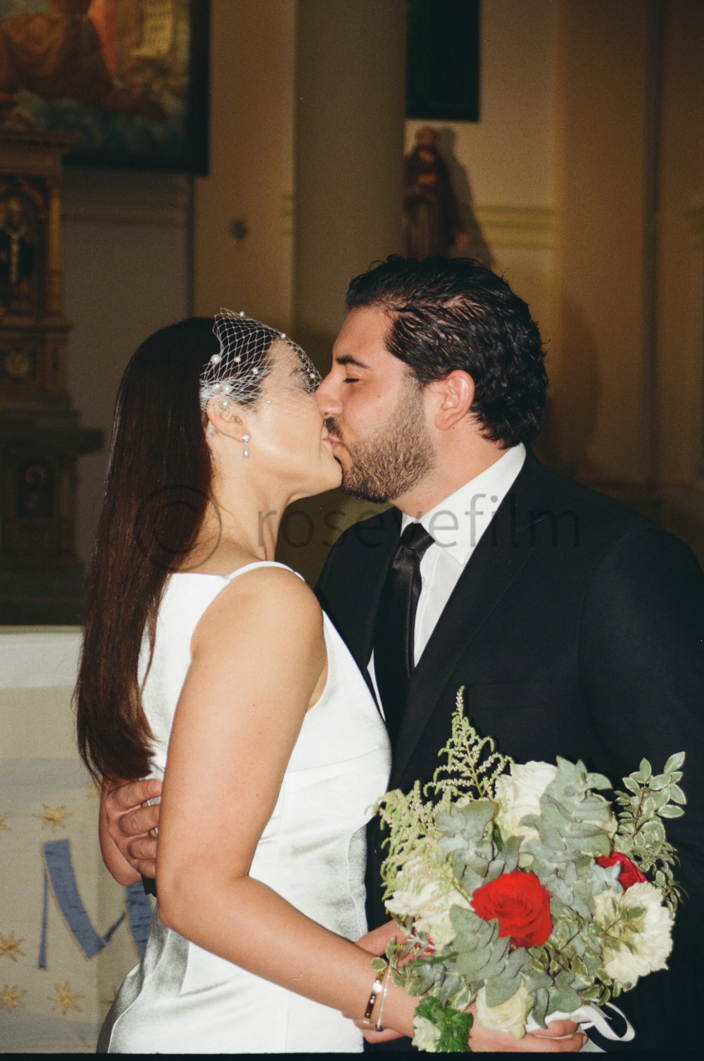 A bride and groom share a kiss inside a church, with the bride holding a bouquet of white and red flowers. The bride wears a white dress and a veil, and the groom is dressed in a black suit with a white shirt and black tie.