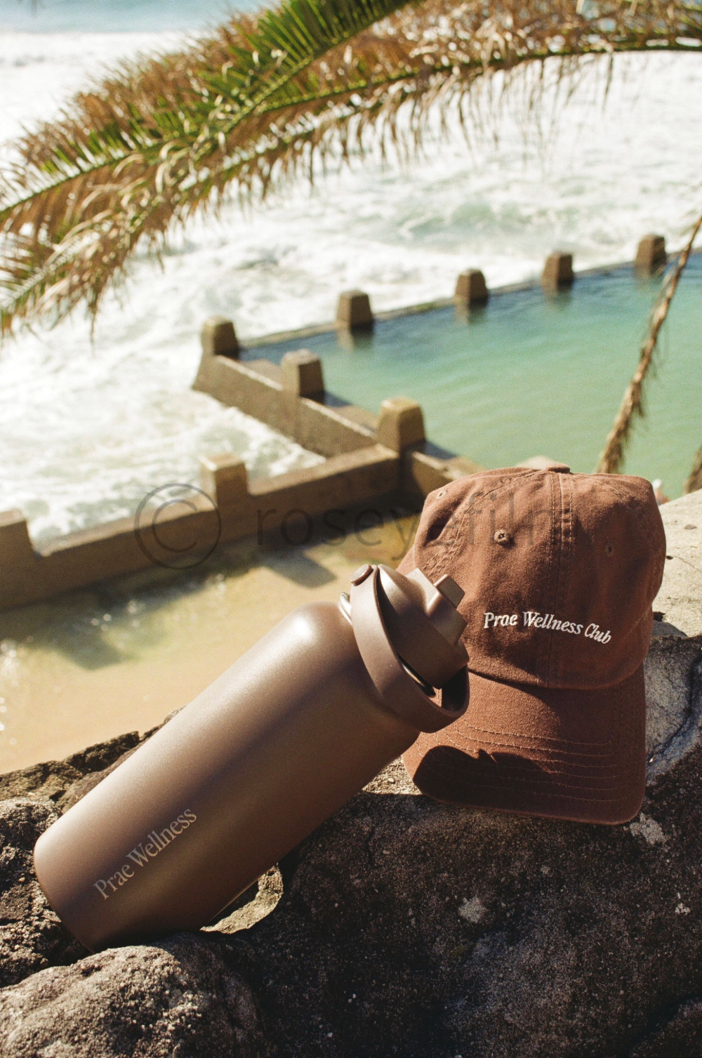 A tan water bottle and a brown cap with 'Prae Wellness Club' embroidered on it, placed on rocks by a water feature with a palm tree over a body of water in the background.