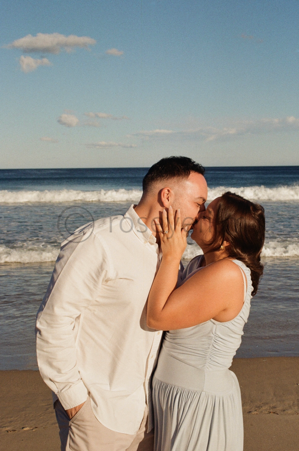 A couple kissing on the beach during sunset, with the ocean and sky in the background.