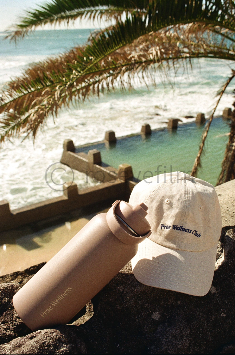 A beige water bottle and a white baseball cap with the embroidered text "Pre Wellness Club" are placed on rocks near a beach, with a wooden pool and ocean waves in the background, framed by palm leaves.