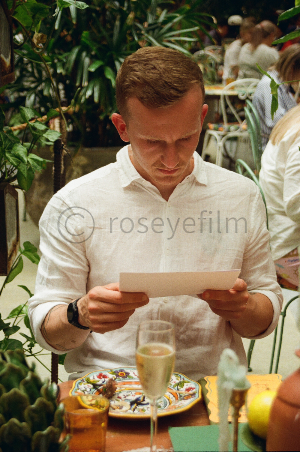 A man in a white shirt looking at a piece of paper in a restaurant with greenery and other diners in the background