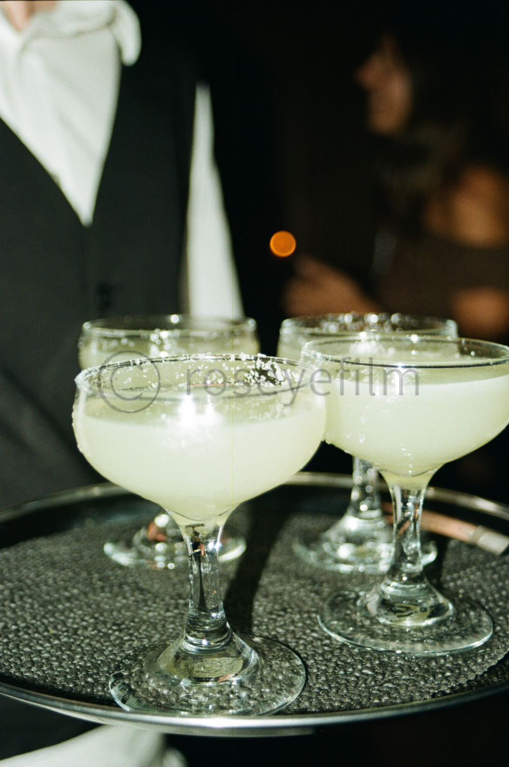 Four coupe glasses filled with a pale yellow cocktail on a tray, with a server in a black suit and white shirt in the background at a social event.
