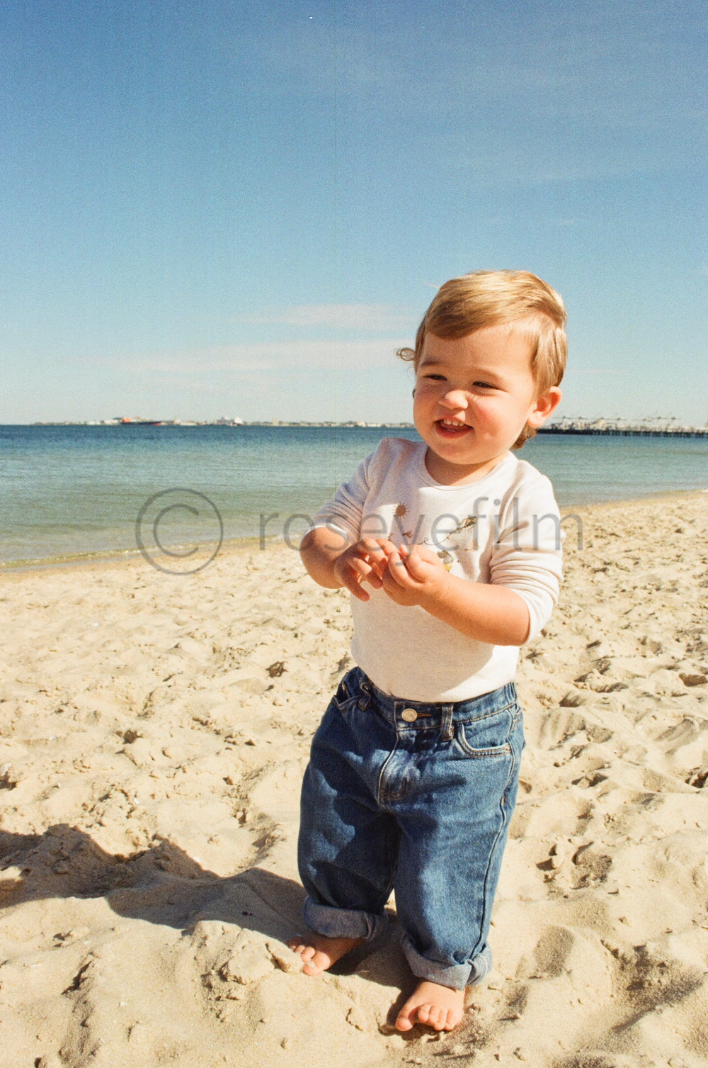A smiling young child with light brown hair standing barefoot on sandy beach near water, wearing a light-colored long sleeve shirt and rolled-up blue jeans.