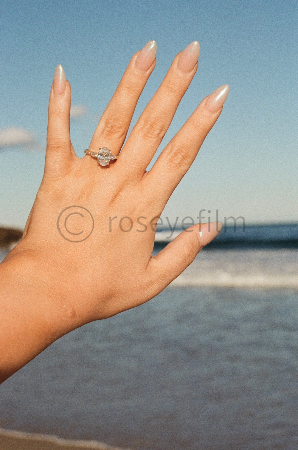 A woman's hand showing an engagement ring with a cluster of diamonds, against a backdrop of a beach and ocean under a partly cloudy sky.