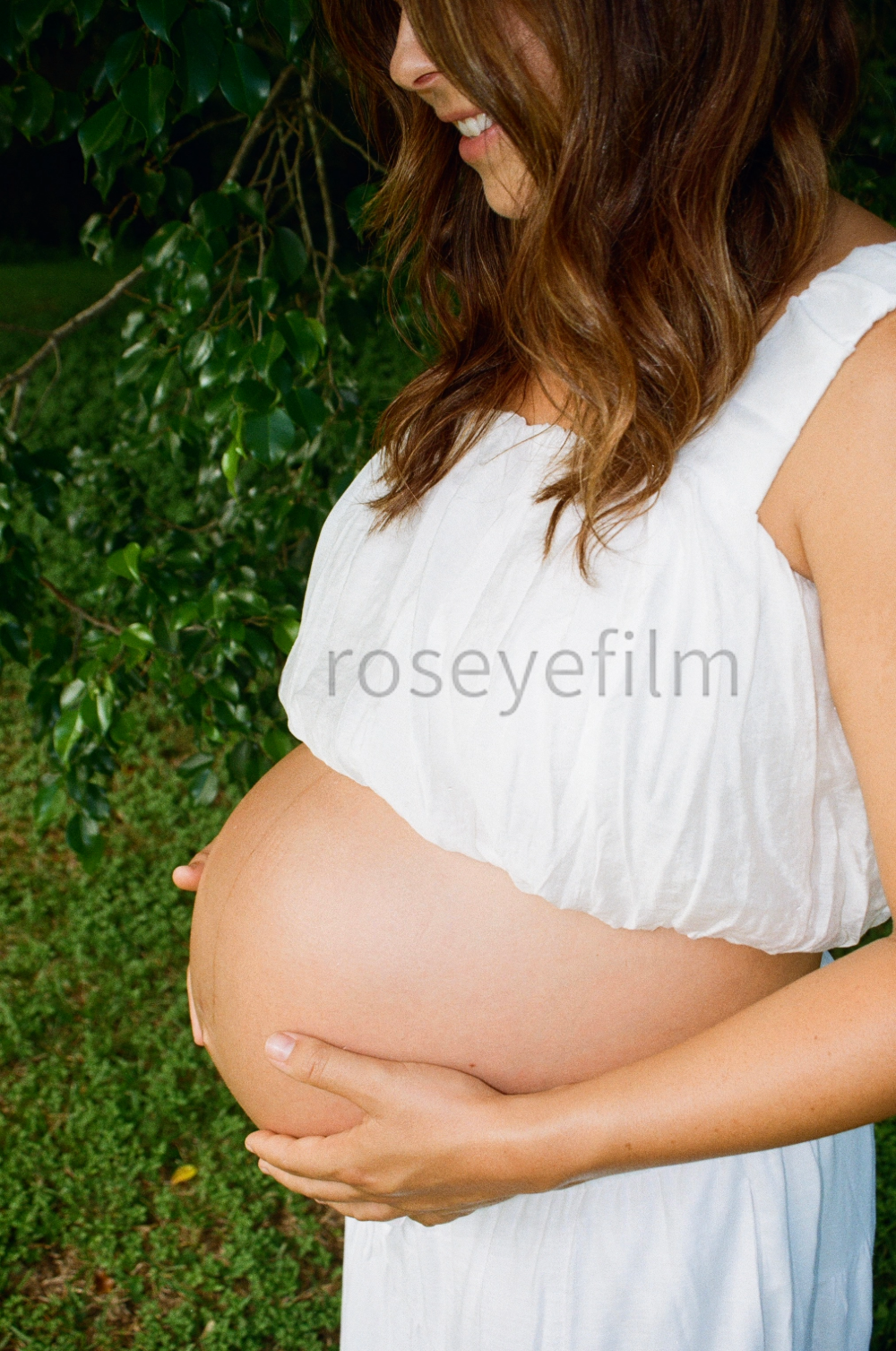 Pregnant woman smiling outdoors, holding her belly with both hands, wearing a white dress with ruffled sleeves, surrounded by greenery.