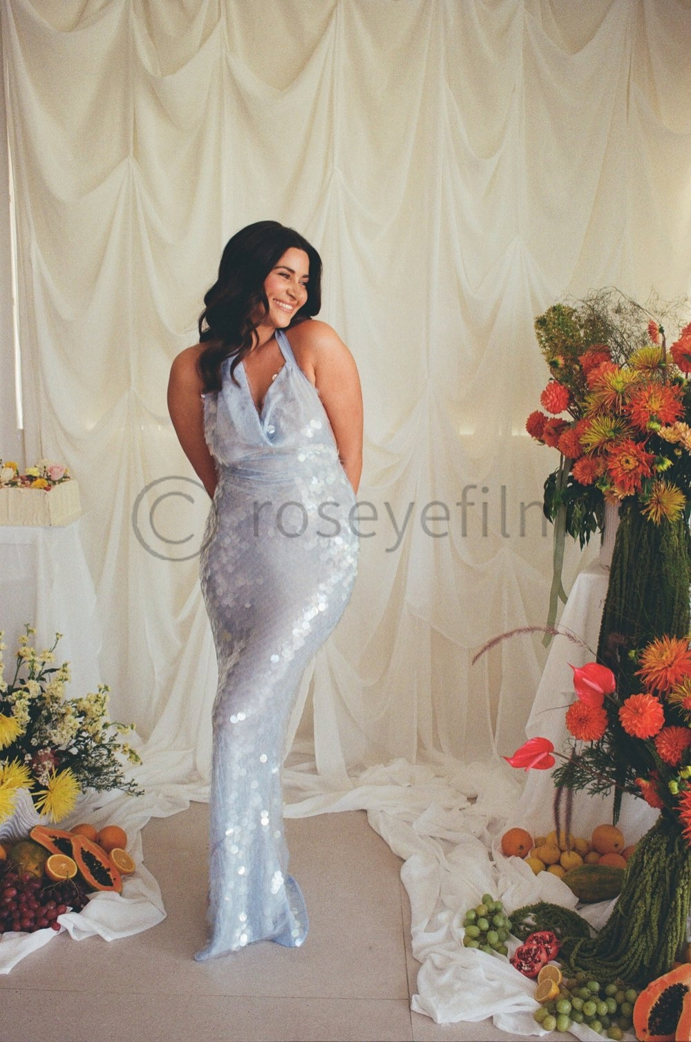 Woman in a sparkling silver dress standing in front of a cream-colored draped backdrop, surrounded by colorful flowers and fruits.