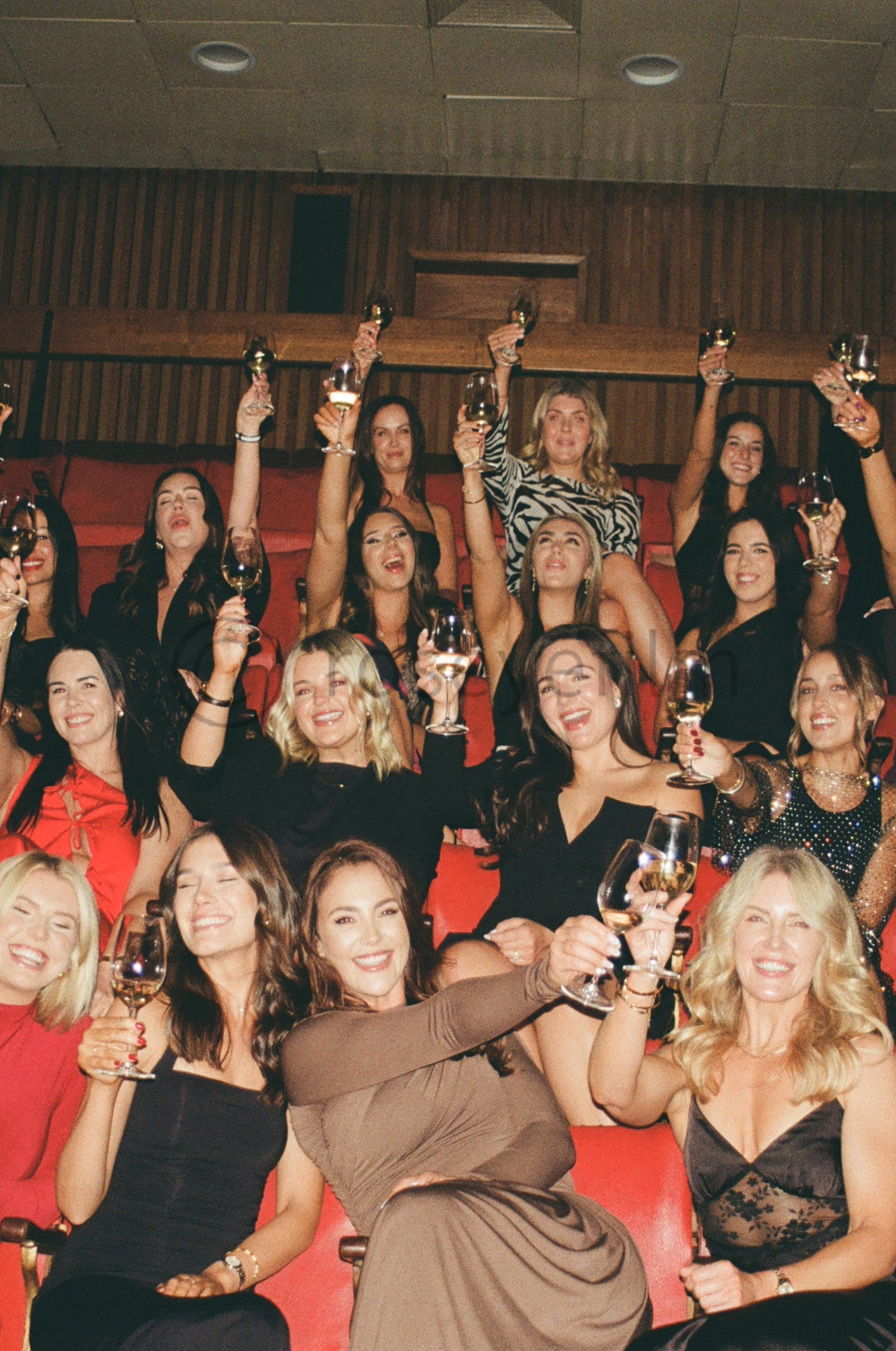 Group of women celebrating, sitting in theater seats, raising glasses of white wine and smiling at the camera.