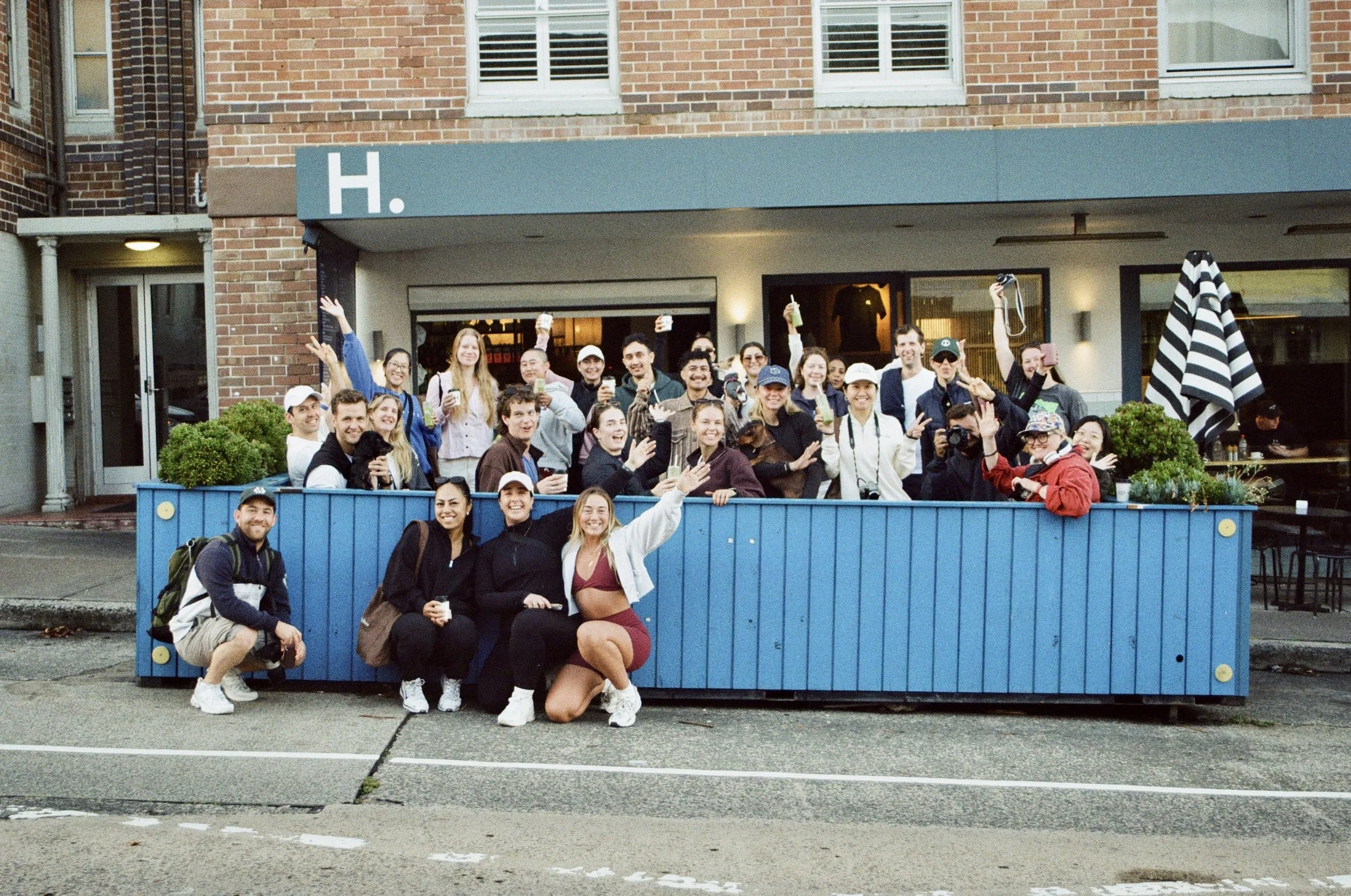 Group of smiling young adults gathered around a blue outdoor railing in front of a building, holding drinks, waving, and taking photos during daytime.