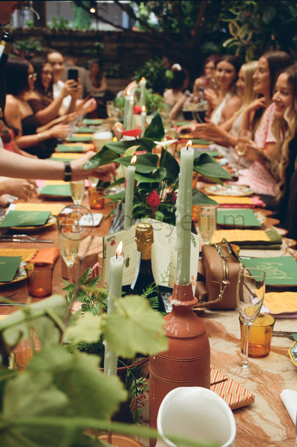 A large outdoor dinner party with many women sitting along a long table decorated with green tablecloth, candles, and foliage. The women are smiling and chatting, some are taking photos with their phones. The table is set with plates, glasses, and co