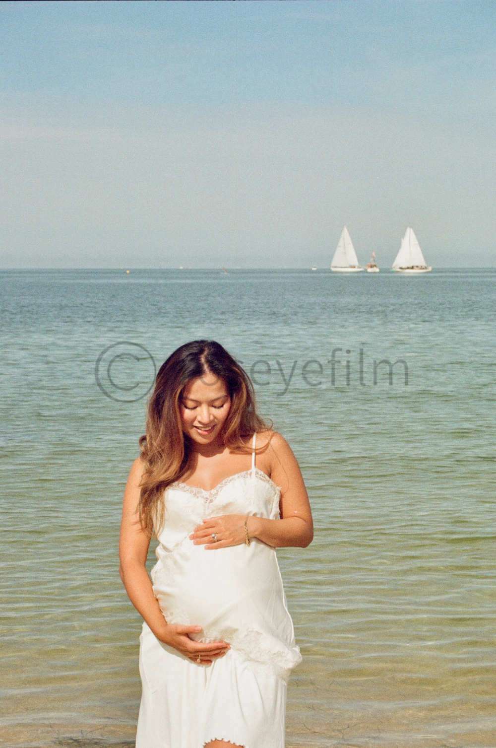 A pregnant woman in a white dress standing on the beach, holding her baby bump with one hand and her belly with the other, smiling and looking down, with the ocean and sailboats in the background.