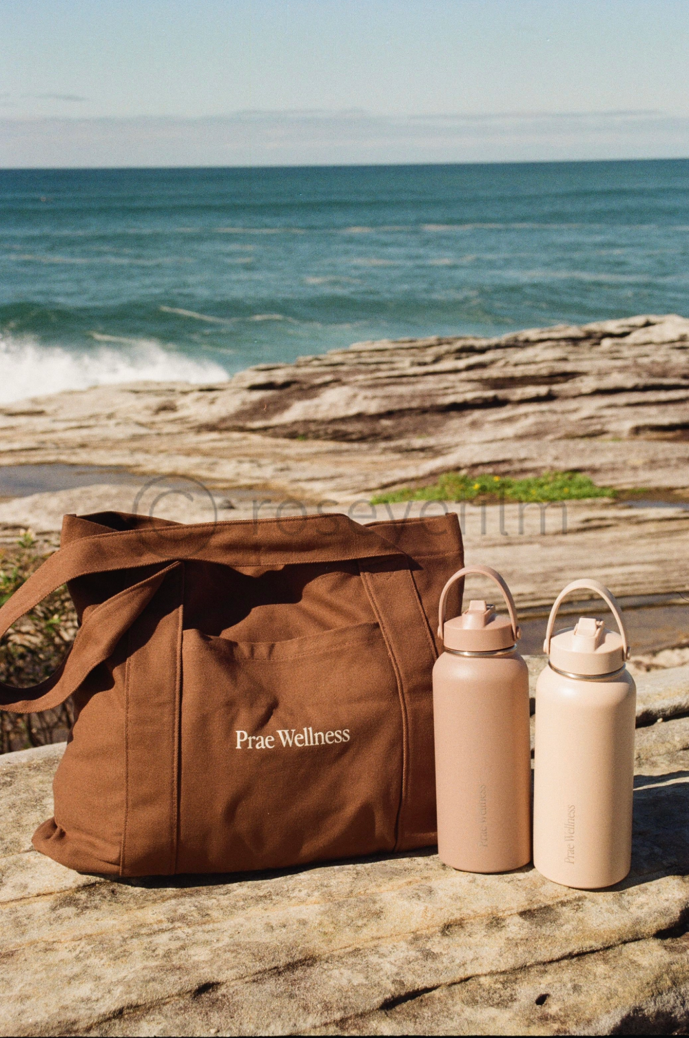 A brown bag labeled 'Prae Wellness' and two matching beige water bottles on a rock by the ocean, with waves and a cloudy sky in the background.