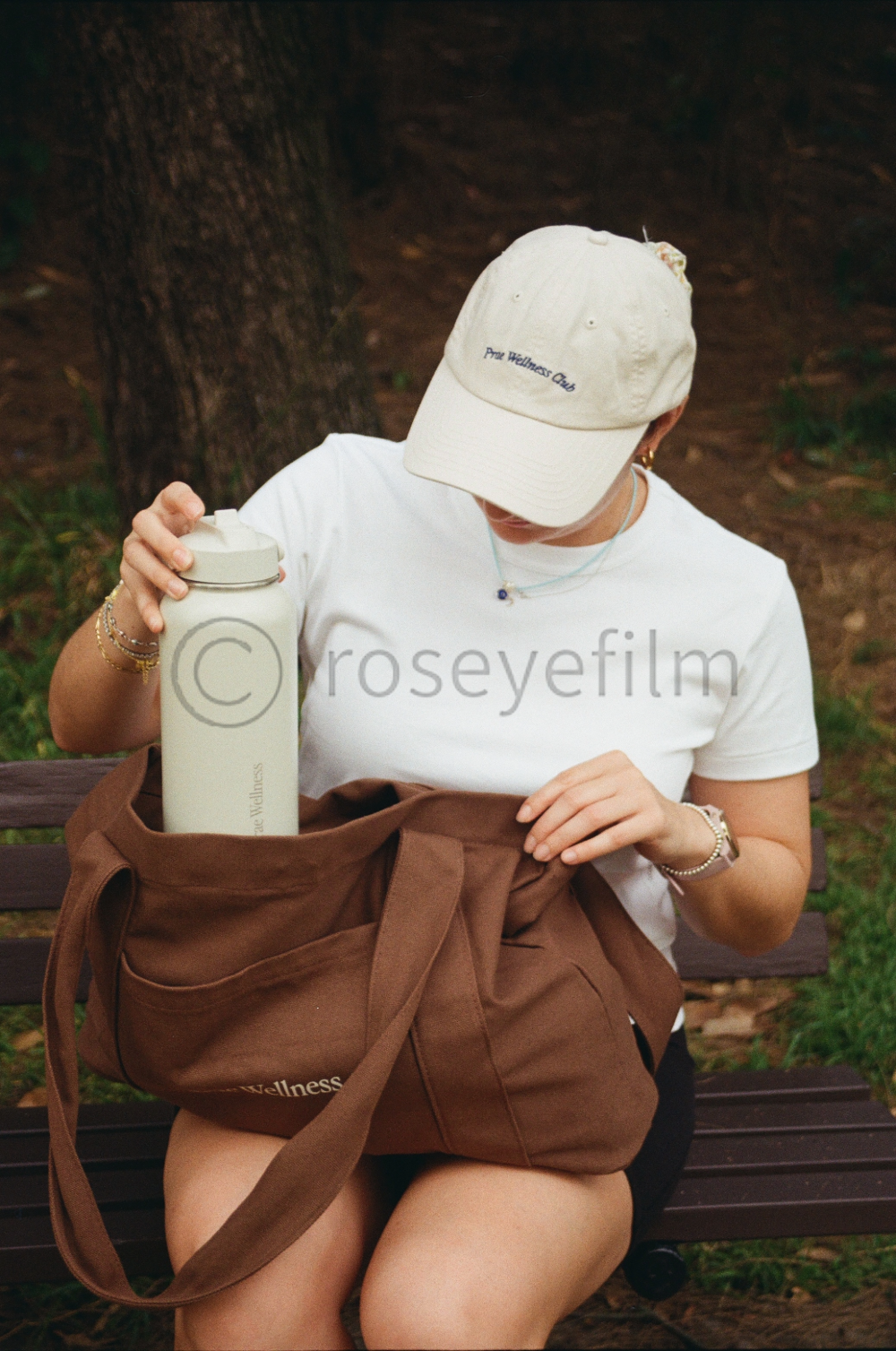 A woman sitting on a park bench, wearing a white cap and T-shirt, reaching into a brown tote bag to grab a cream-colored water bottle.