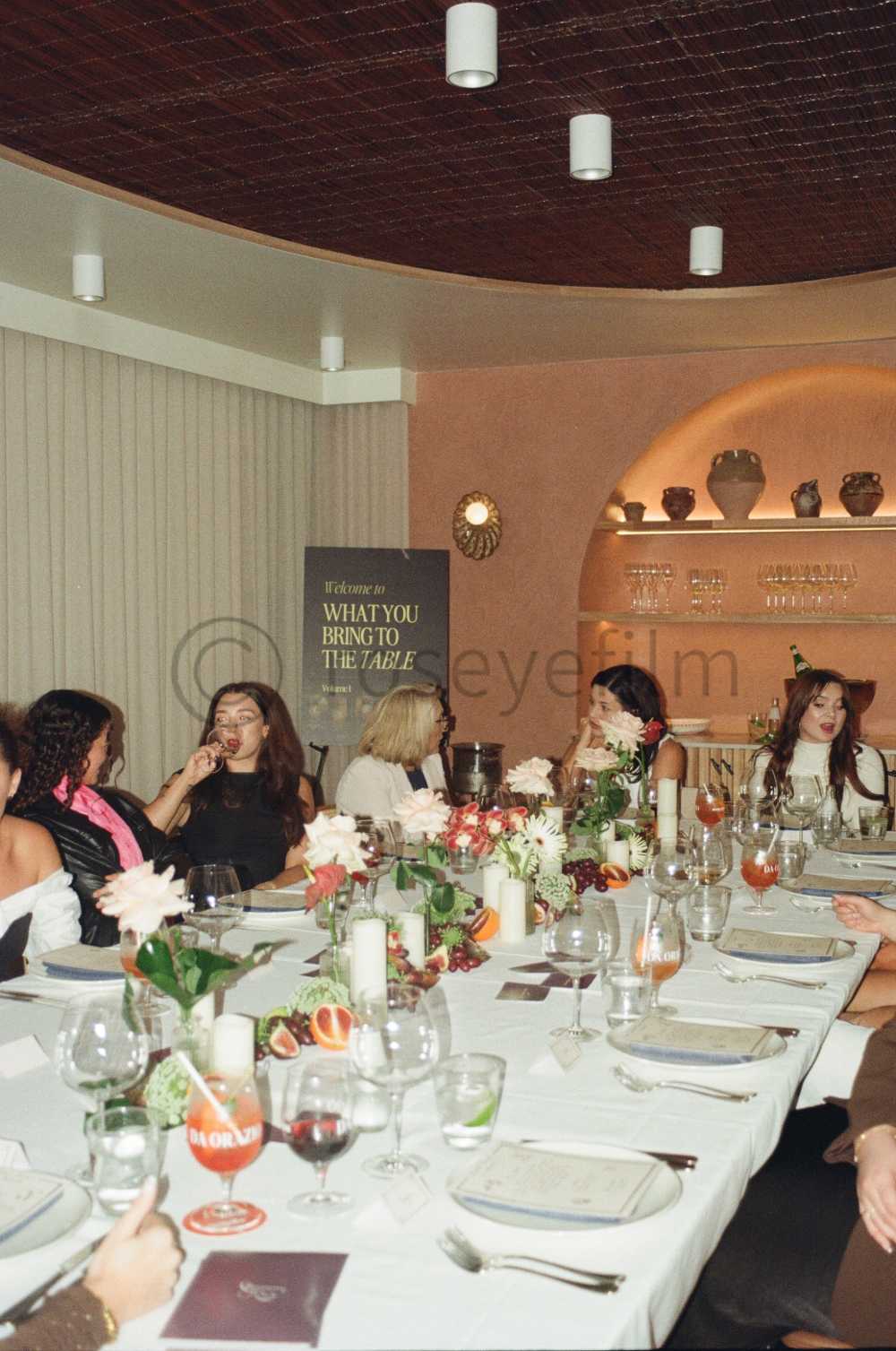 A group of women sitting around a long dining table decorated with flowers, candles, and fruit at an elegant restaurant or event space.