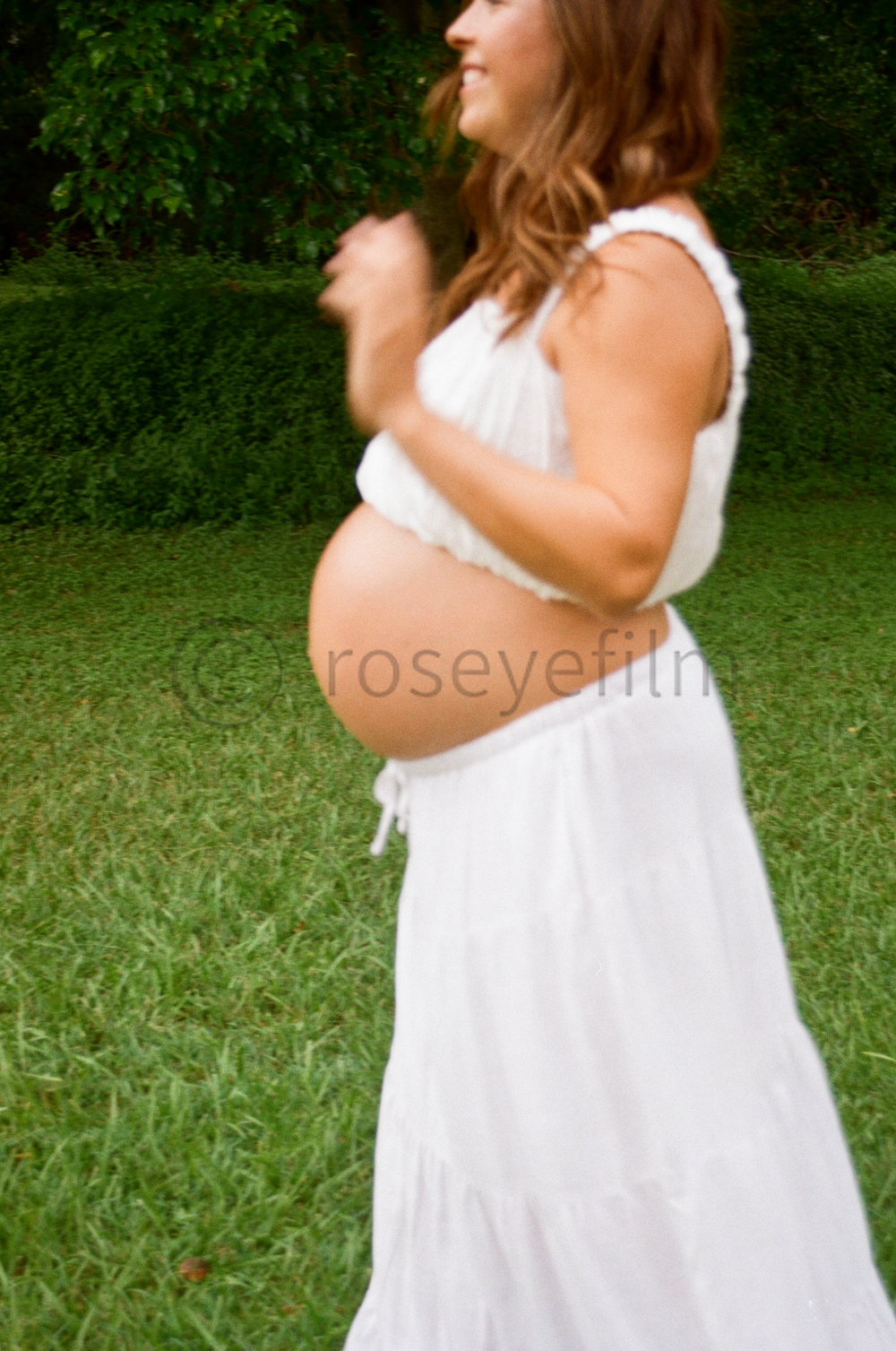 Pregnant woman outdoors wearing a white sleeveless top and skirt, with green foliage in the background.