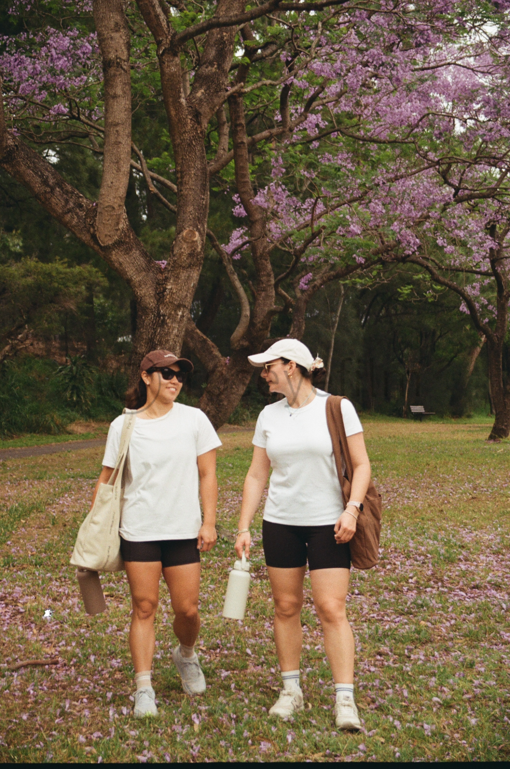 Two women walking in a park with purple flowering trees in the background, dressed in casual athletic wear, one with a beige bag and the other with a brown backpack.