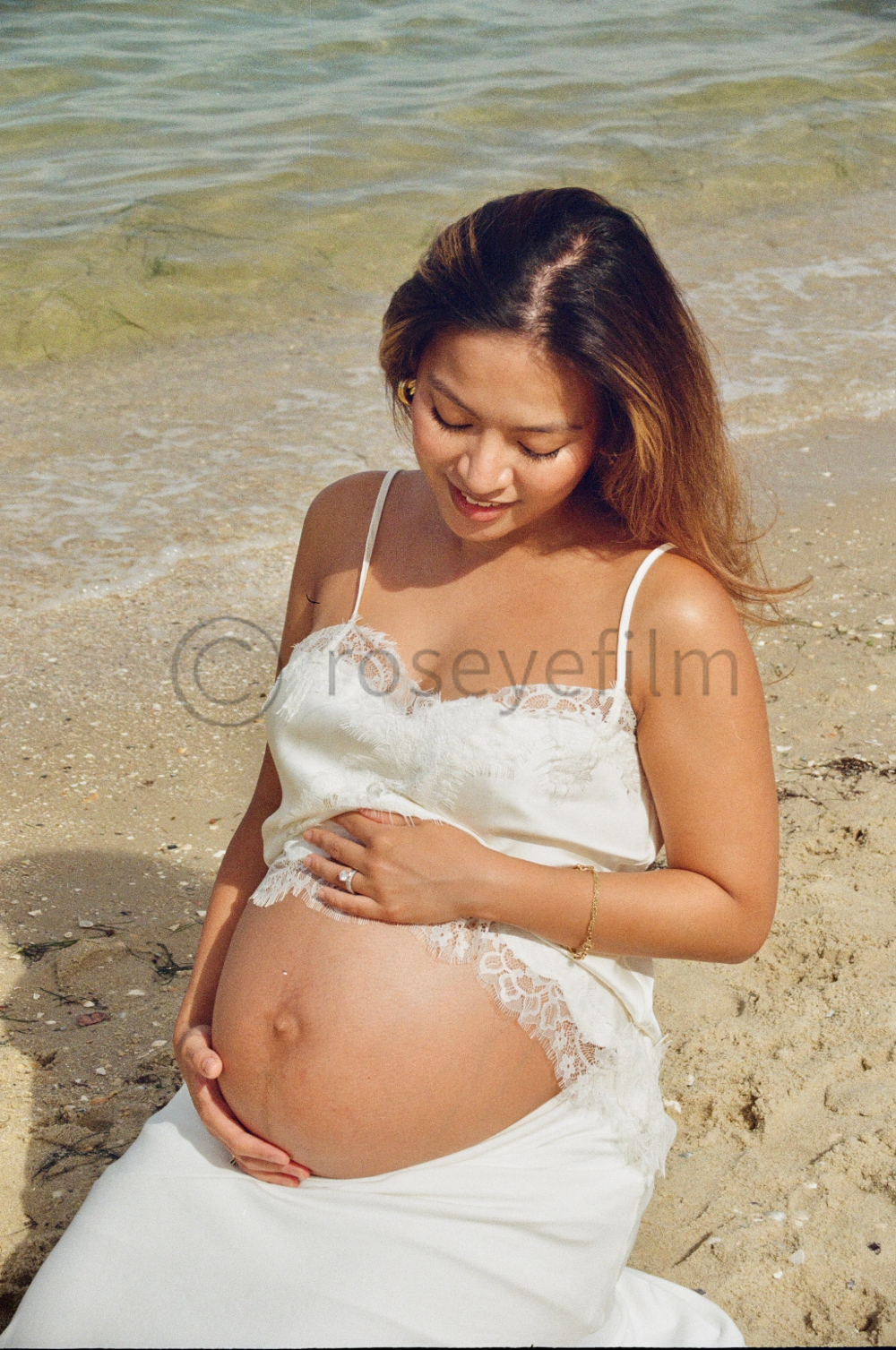 Pregnant woman in white dress sitting on sandy beach, holding her belly and smiling, with ocean waves in the background.