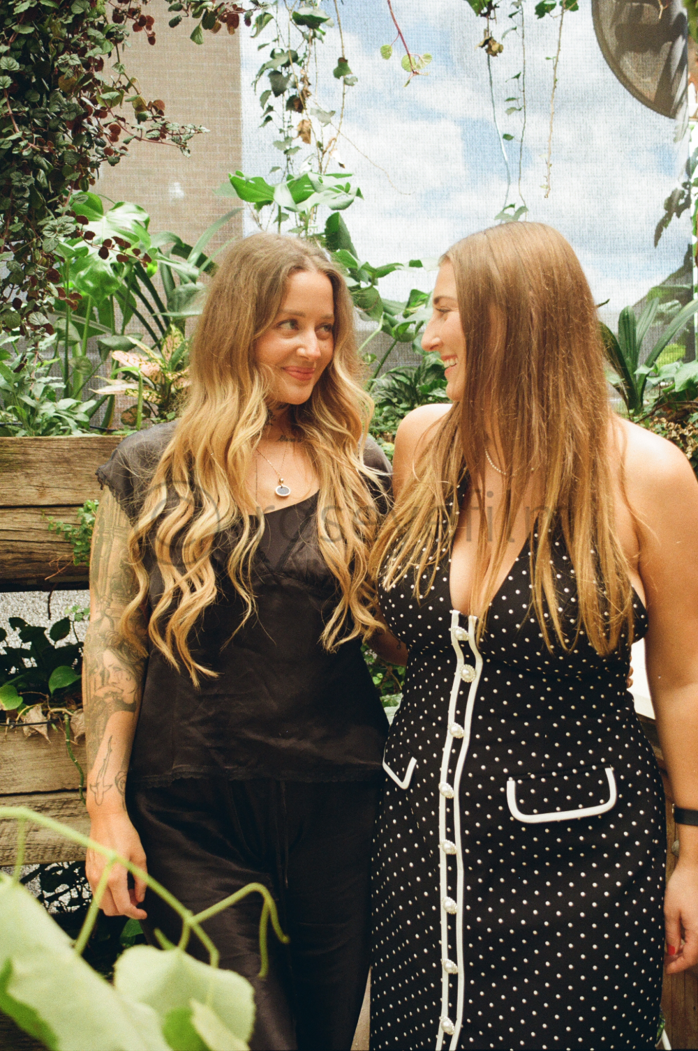 Two women are standing close together, smiling at each other in a lush indoor garden with green plants and a wooden structure in the background.