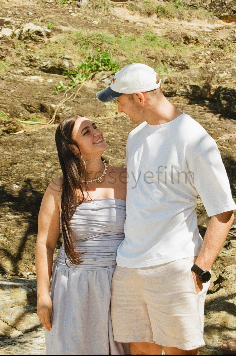 A happy couple standing close together outdoors on a rocky surface, smiling and gazing at each other.