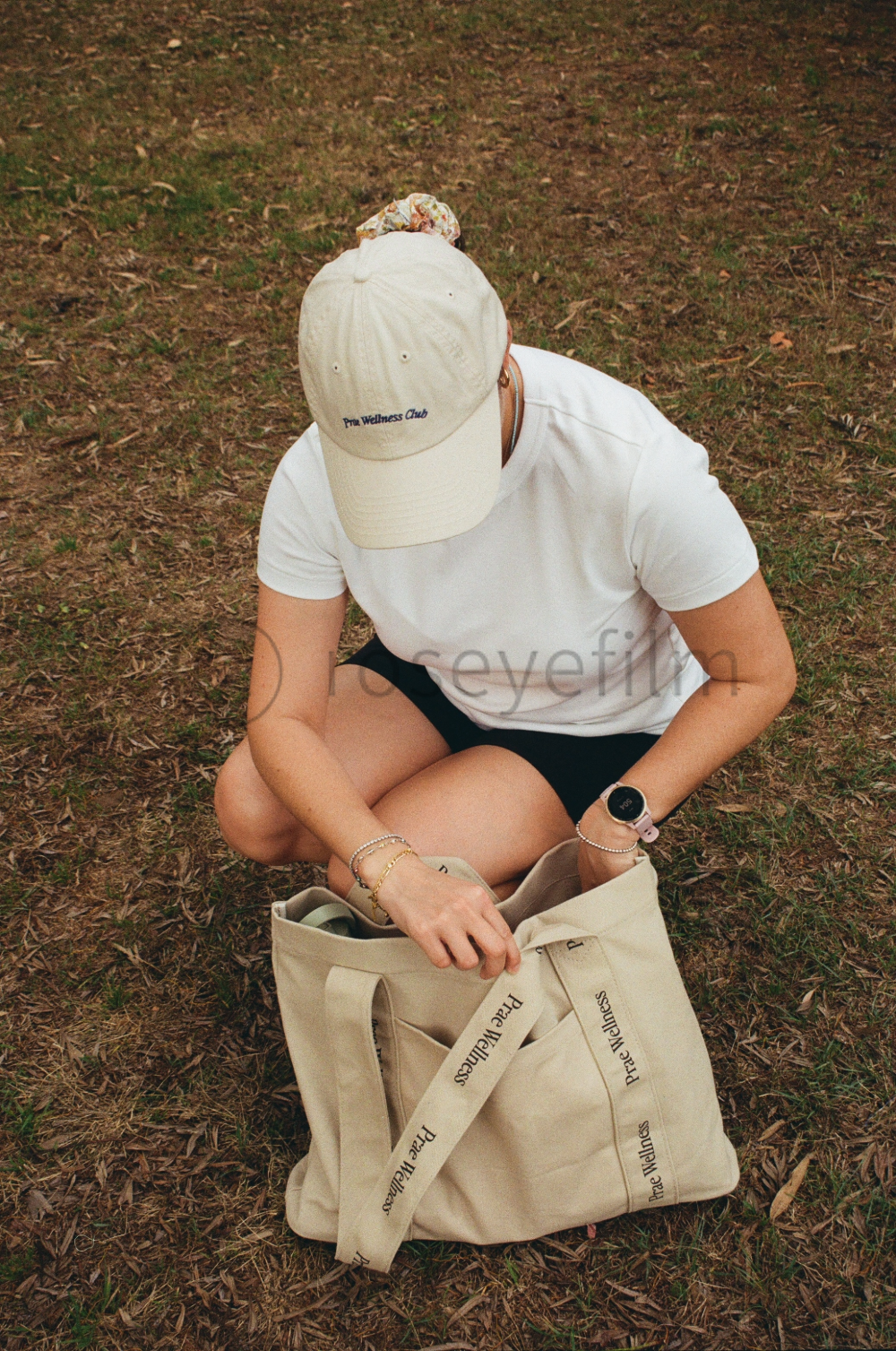 A person sitting on the ground outdoors, wearing a beige cap, a white t-shirt, and black shorts, packing a large beige tote bag with 'Pree Wellness' written on it.