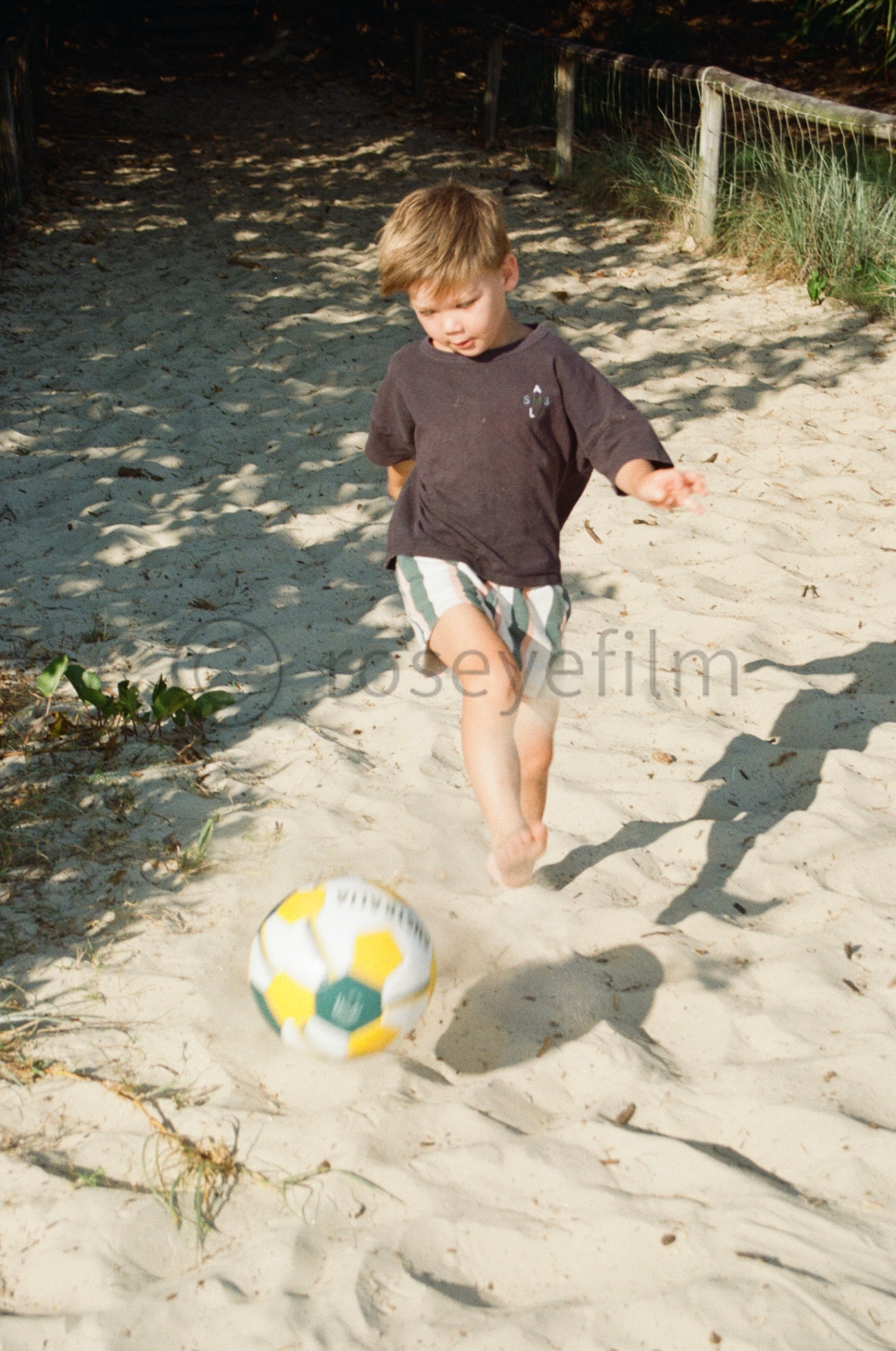 A young boy with light brown hair playing soccer on a sandy beach. He's barefoot, wearing a dark T-shirt and striped shorts, and is kicking a yellow and white soccer ball as he runs.