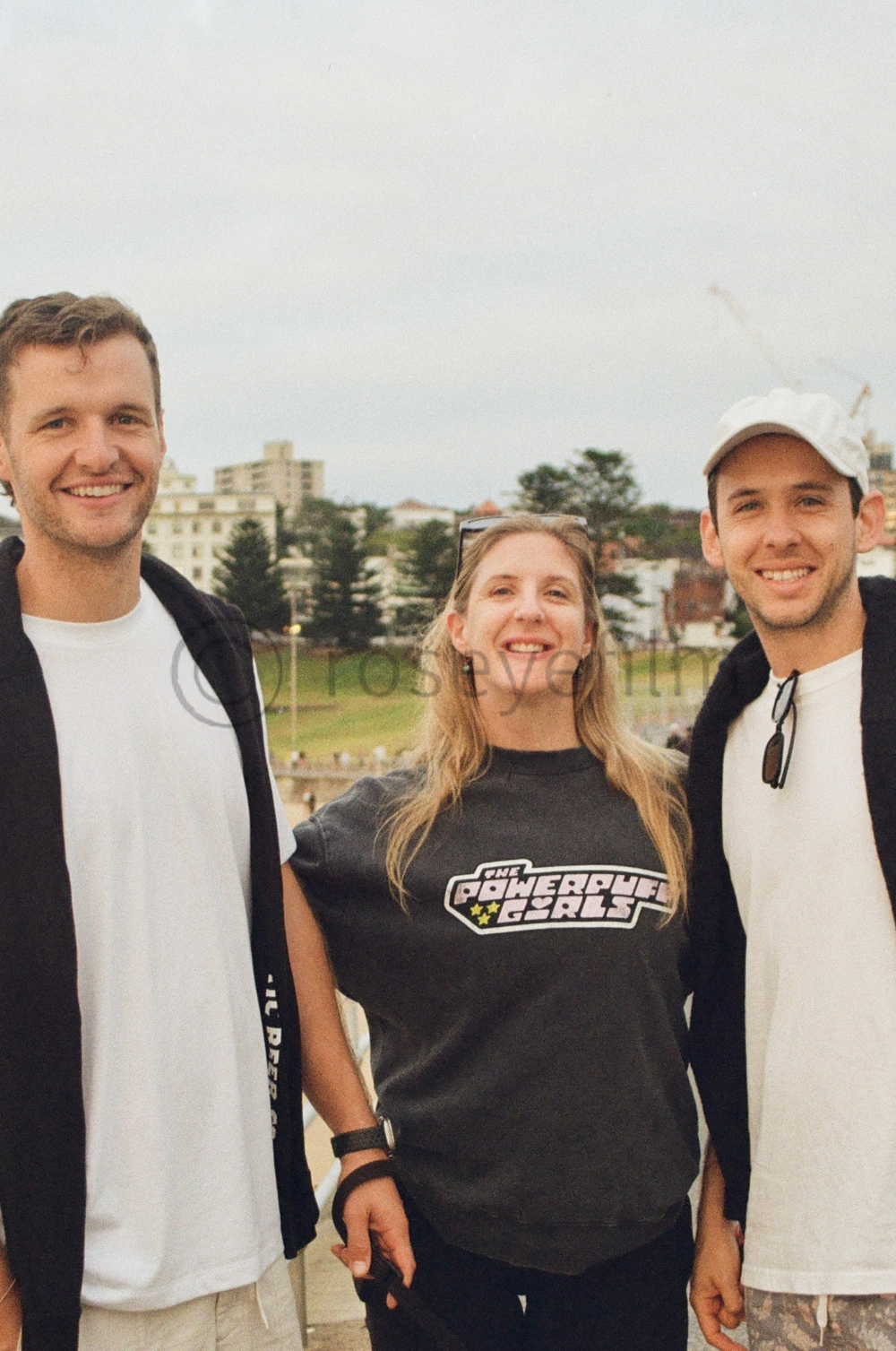 Three young adults smiling outdoors in a park or field, with buildings and trees in the background. The woman in the middle is wearing a black T-shirt with "The Powercruise" logo, and the man on the right is wearing a white cap and sunglasses hanging