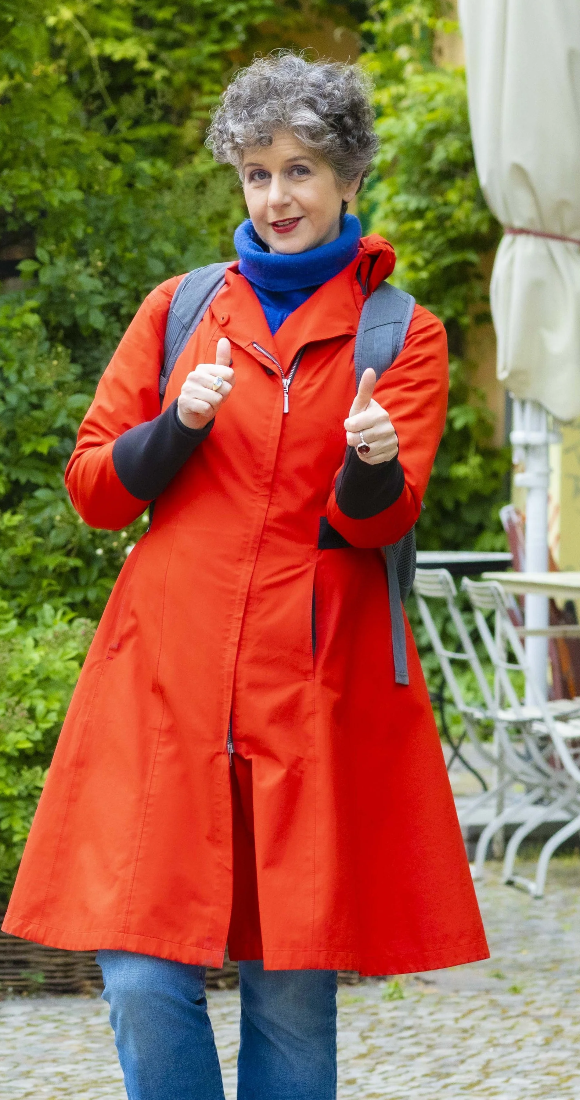 Shlomit giving two thumbs up, wearing a red raincoat, blue scarf, and jeans, standing outdoors with trees and outdoor furniture in the background.