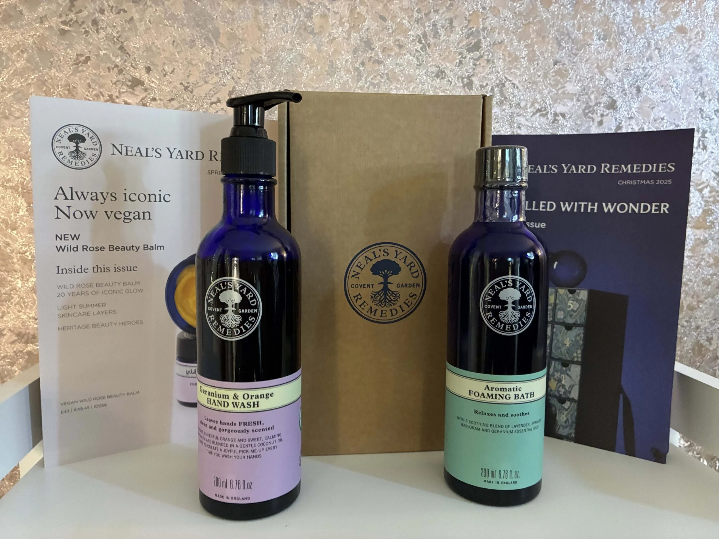 Two Neal's Yard Remedies bottles, one labeled Geranium & Orange Hand Wash and the other Aromatic Foaming Bath, with informational leaflets and a brown box in the background.