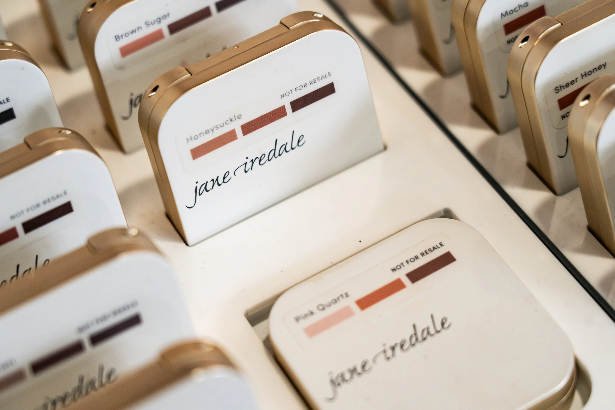Multiple makeup blush compact cases labeled with shades such as Honey Suckle, Pink Quartz, Mocha, Sheer Honey, Brown Sugar, and other shades, stacked on a display shelf.