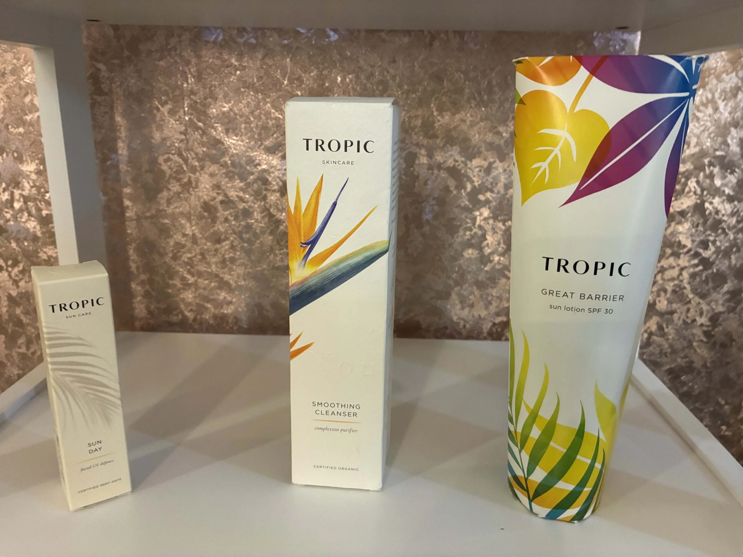 Tropic skincare products including a small box labeled 'Sun Care - Sun Day,' a larger box labeled 'Smoothing Cleanser,' and a tube labeled 'Great Barrier Sun Lotion SPF 30,' with colorful tropical leaf and flower illustrations.
