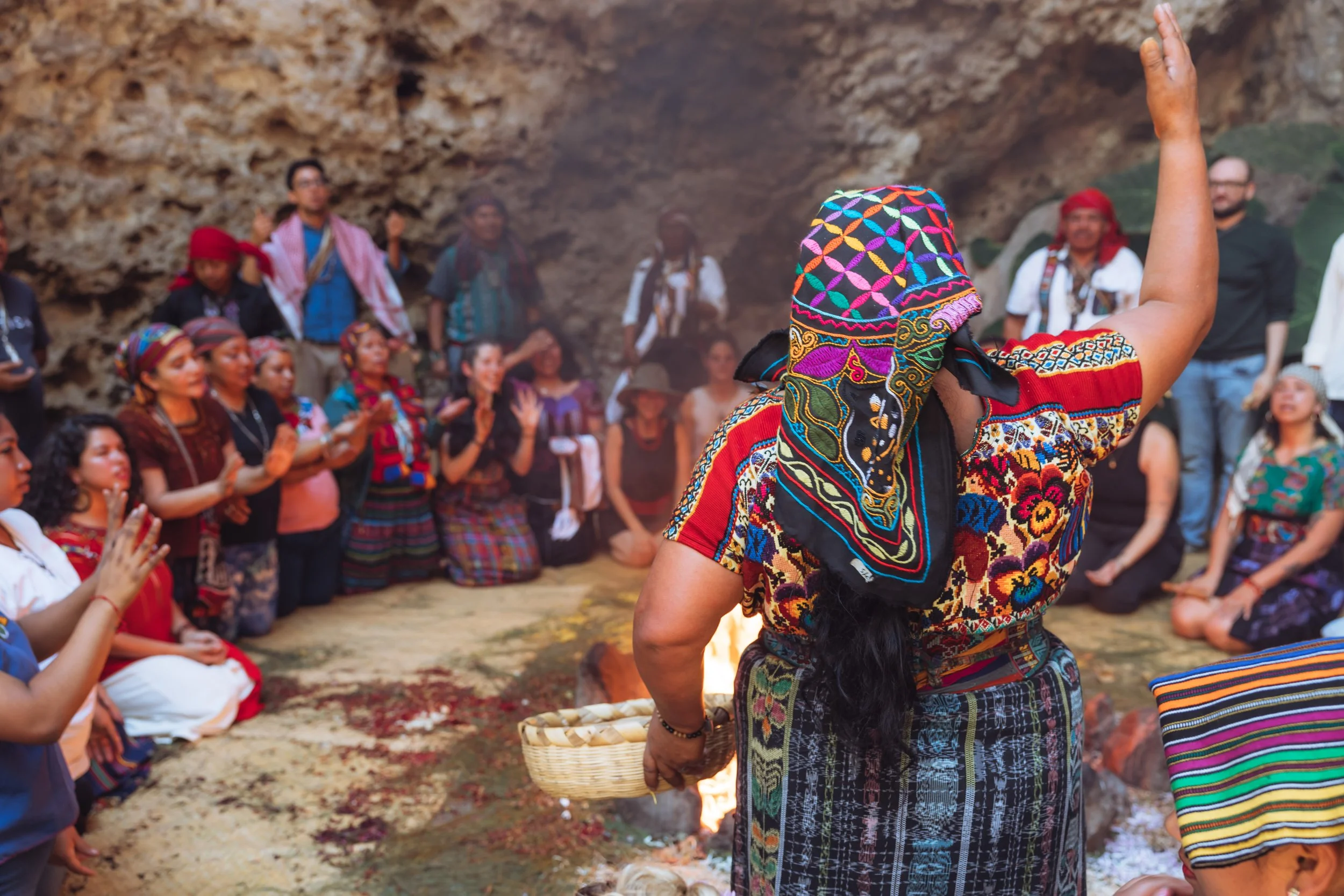 A woman in traditional indigenous clothing performing a ritual around a fire with a group of people sitting and standing around her, inside a cave.