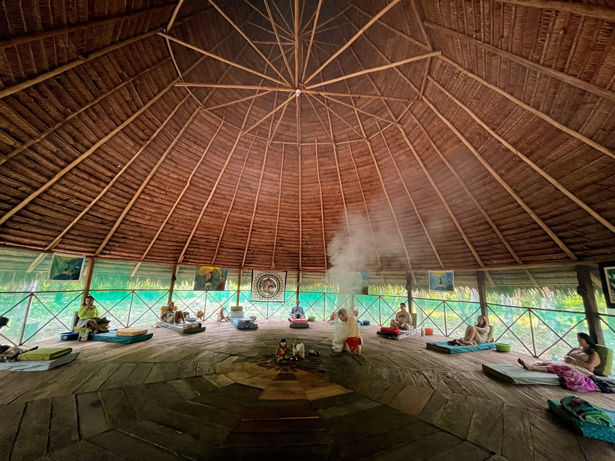 Four individuals sitting on mats in a circular open-air hut with a thatched roof, engaged in a spiritual or meditation session, with smoke rising from a central fire or incense and vibrant artwork on the walls.