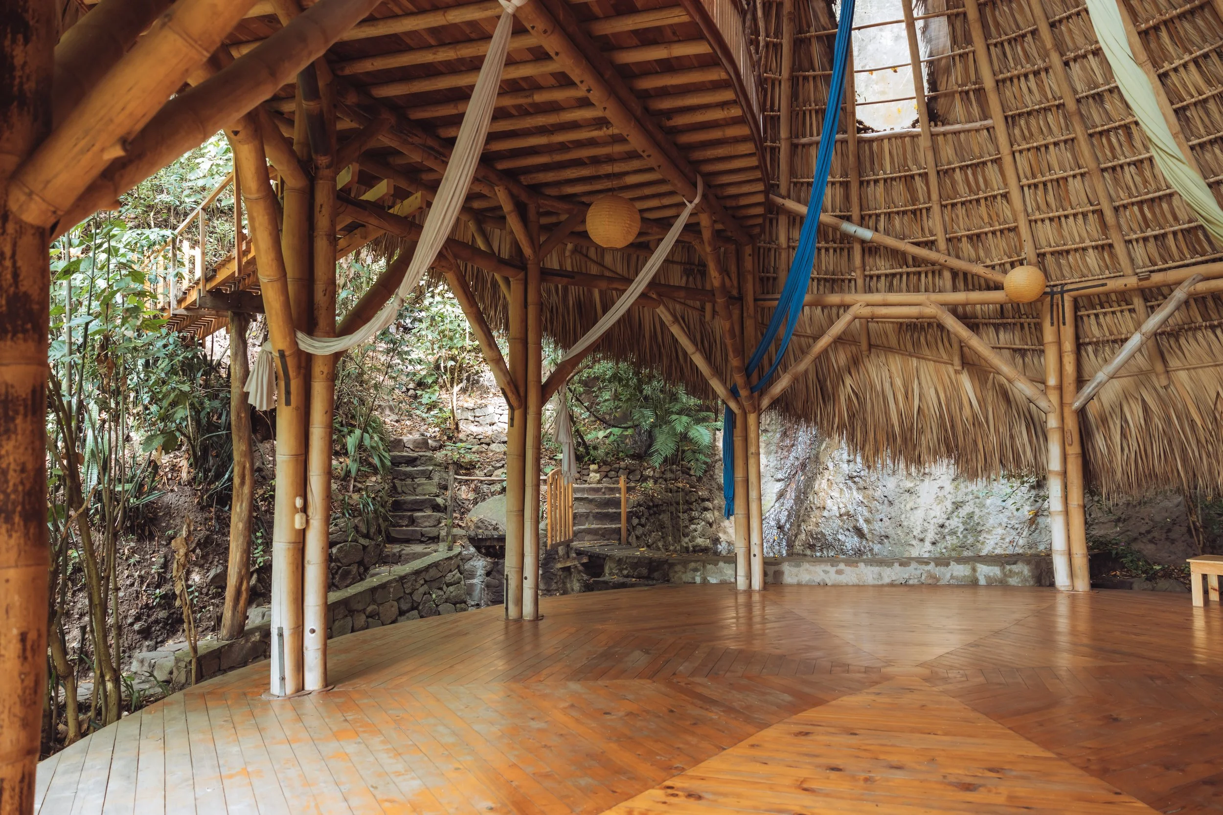 Interior of a rustic open-air hut with bamboo and thatch construction, featuring a wooden floor, hanging fabric, and surrounding greenery with stone steps outside.