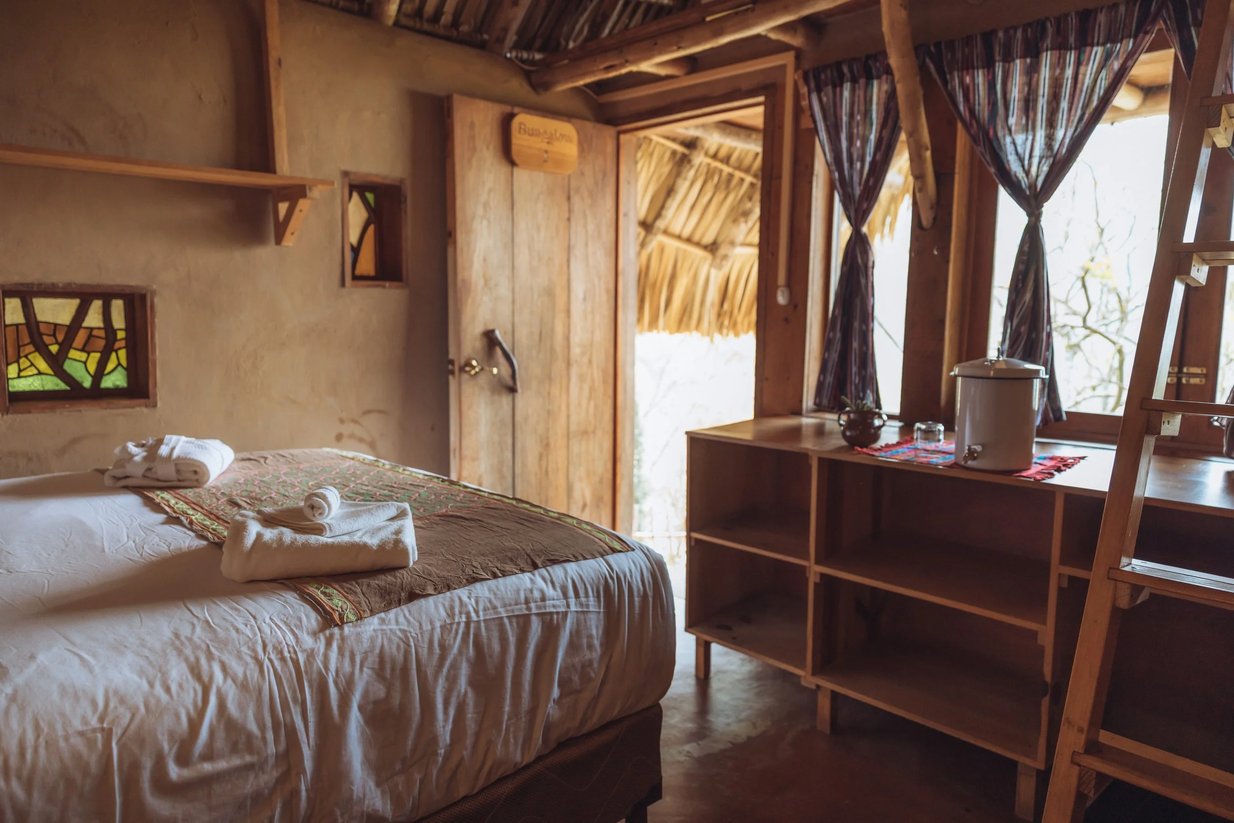 Interior of a rustic bedroom with a neatly made bed, folded towels, wooden shelves, stained glass windows, and an open door revealing a thatched roof outside.